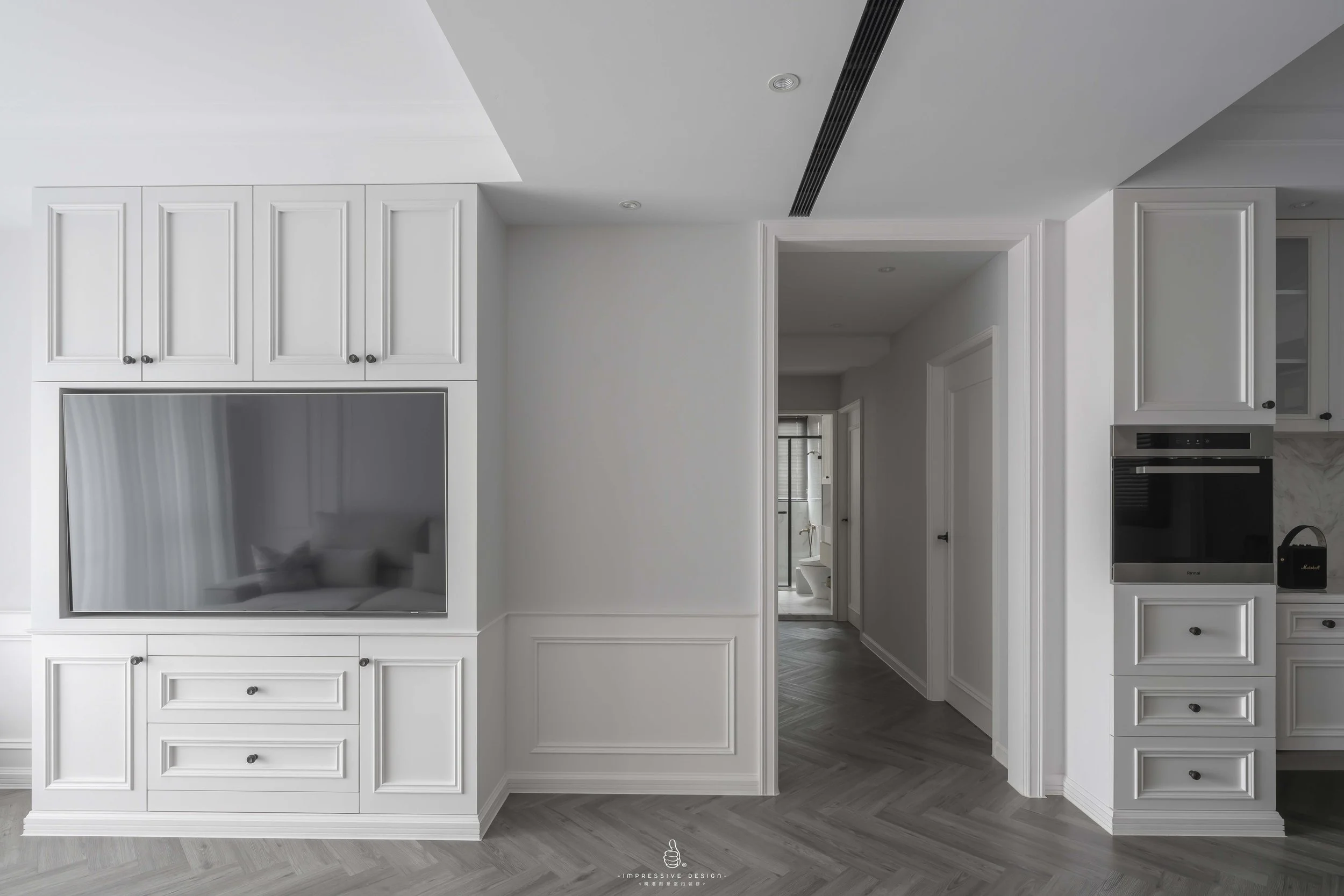 Modern modern white kitchen with built-in appliances and white cabinets. There is a hallway leading to a bathroom with a glass door. The flooring is gray herringbone tile.