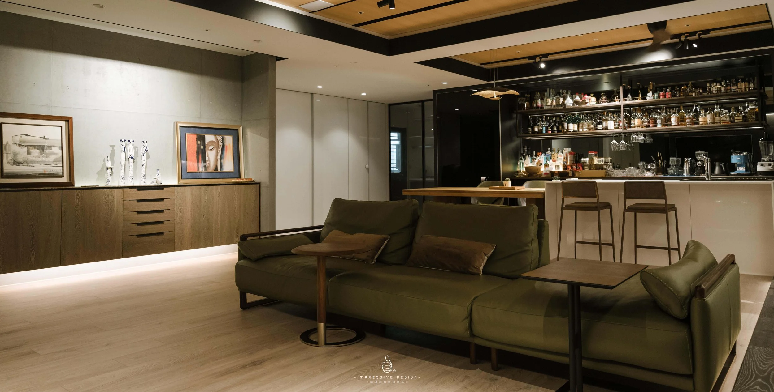 A modern living room with a green sofa, side tables, artwork on the wall, a bar area with bottles and glasses, and bar stools, illuminated by warm lighting.