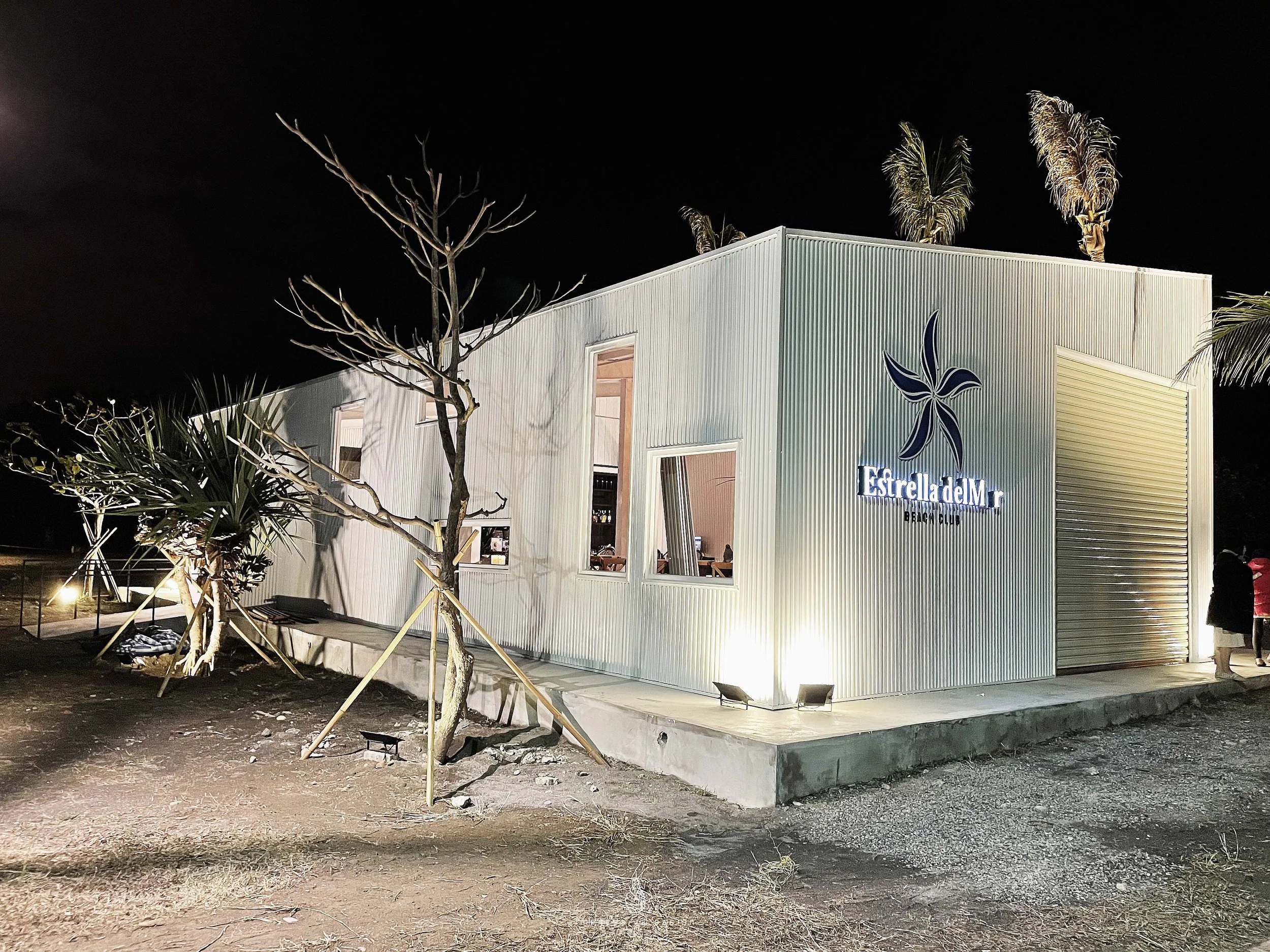 Night view of a modern building with a sign that reads 'Estrella del Mar Beach Club,' illuminated by exterior lights. The building has a metal corrugated exterior and several open windows, with some tall palm trees and a leafless tree nearby.