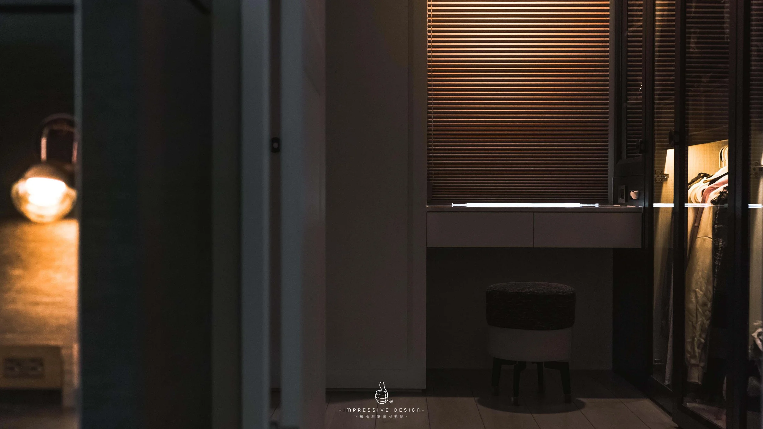 A dimly lit room with a small round stool, closed wooden blinds, and a wardrobe with clothes inside, part of a bed is visible on the right.