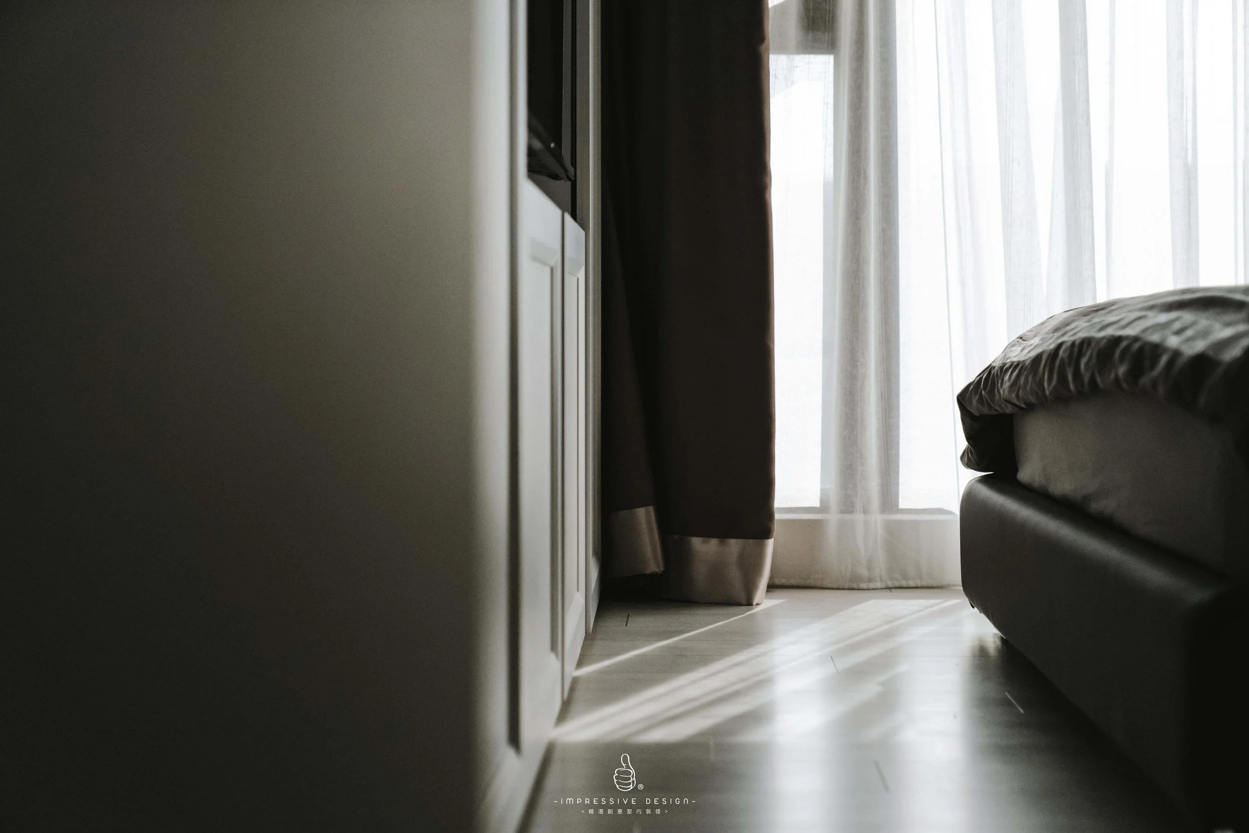 A bedroom with a bed on the right, a window with sheer curtains, a brown curtain, and sunlight streaming through the window, illuminating the floor.