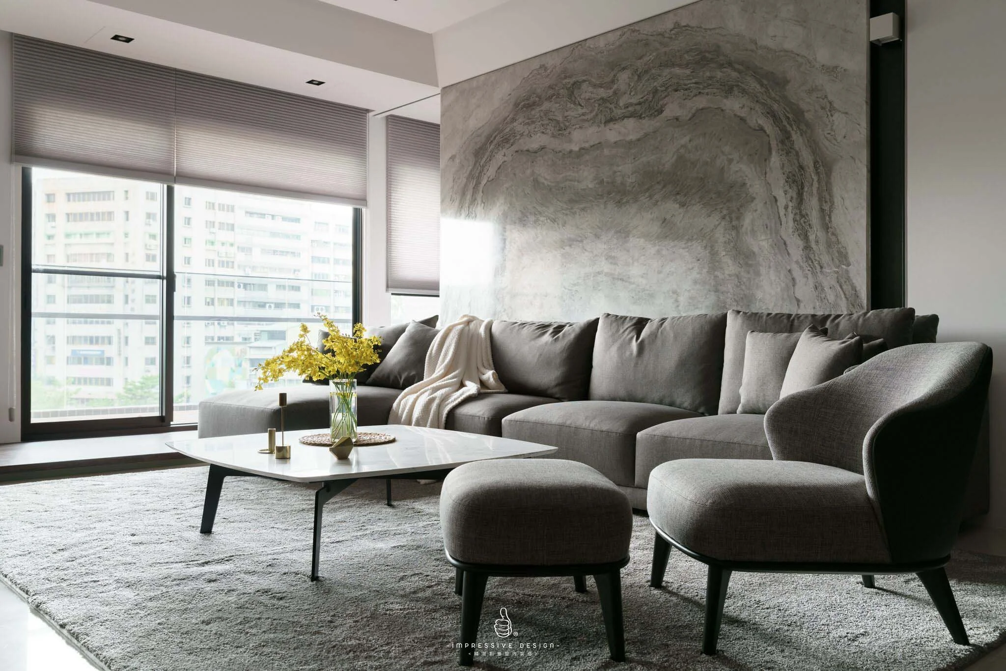 Modern living room with large windows, a gray sectional sofa covered with a white blanket, and a matching armchair with an ottoman. A coffee table with a vase of yellow flowers and decorative objects sits on a textured gray rug. The room features a g