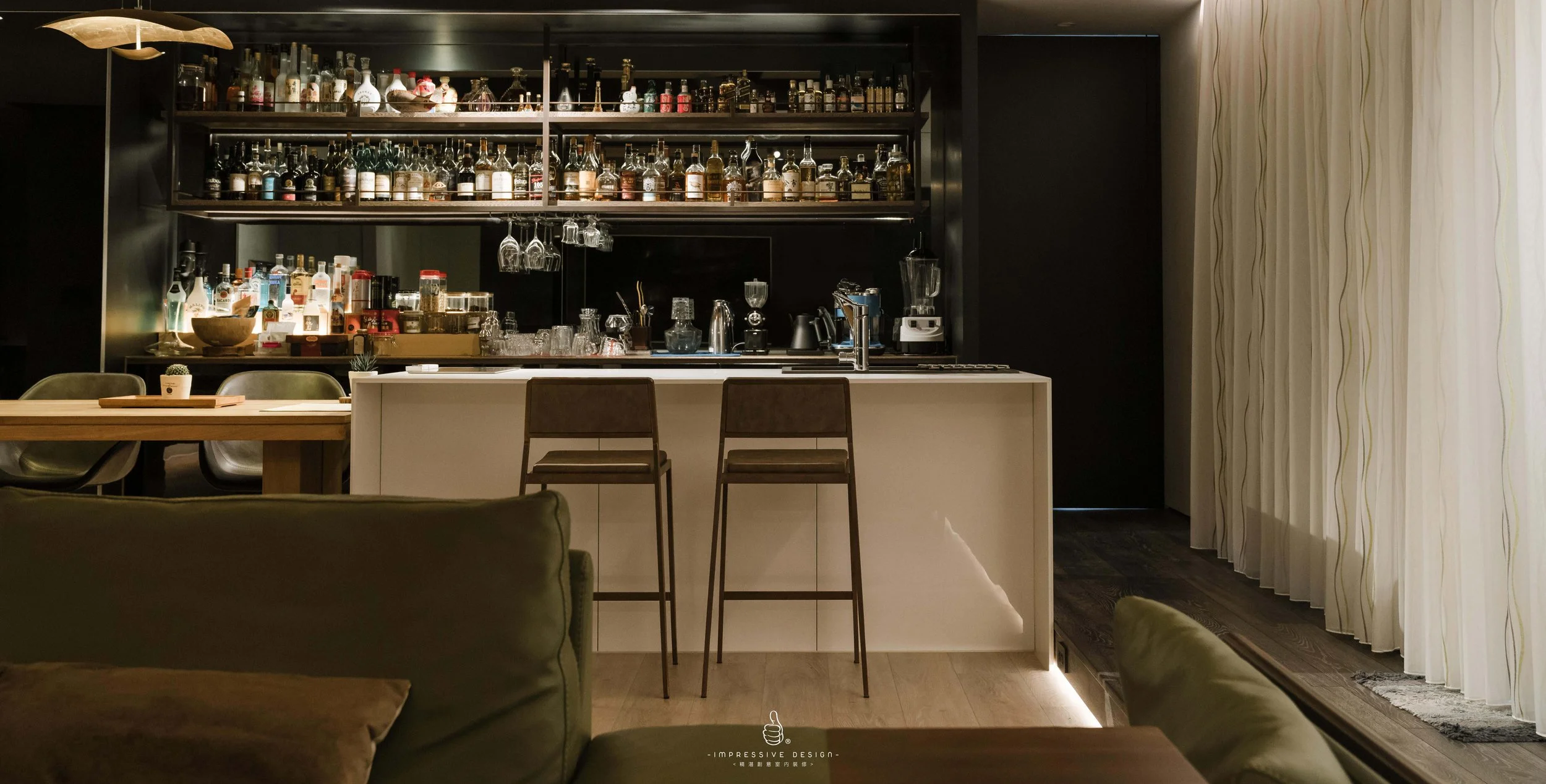 A modern home bar area with a white island counter and two bar stools. Behind the counter are black shelves filled with various bottles of liquor and glassware. To the right, there are large windows with white curtains, allowing natural light into th