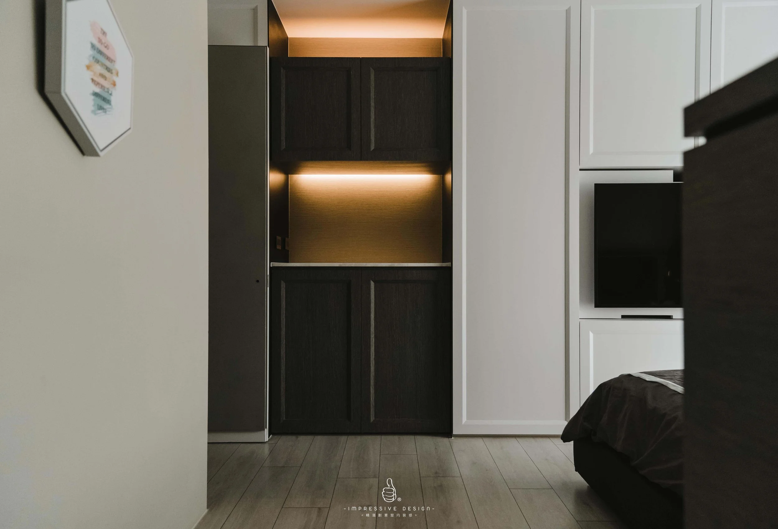 A modern bedroom corner with white and black cabinets, a partially visible bed with dark bedding, a small TV, and a framed wall art on a white wall.