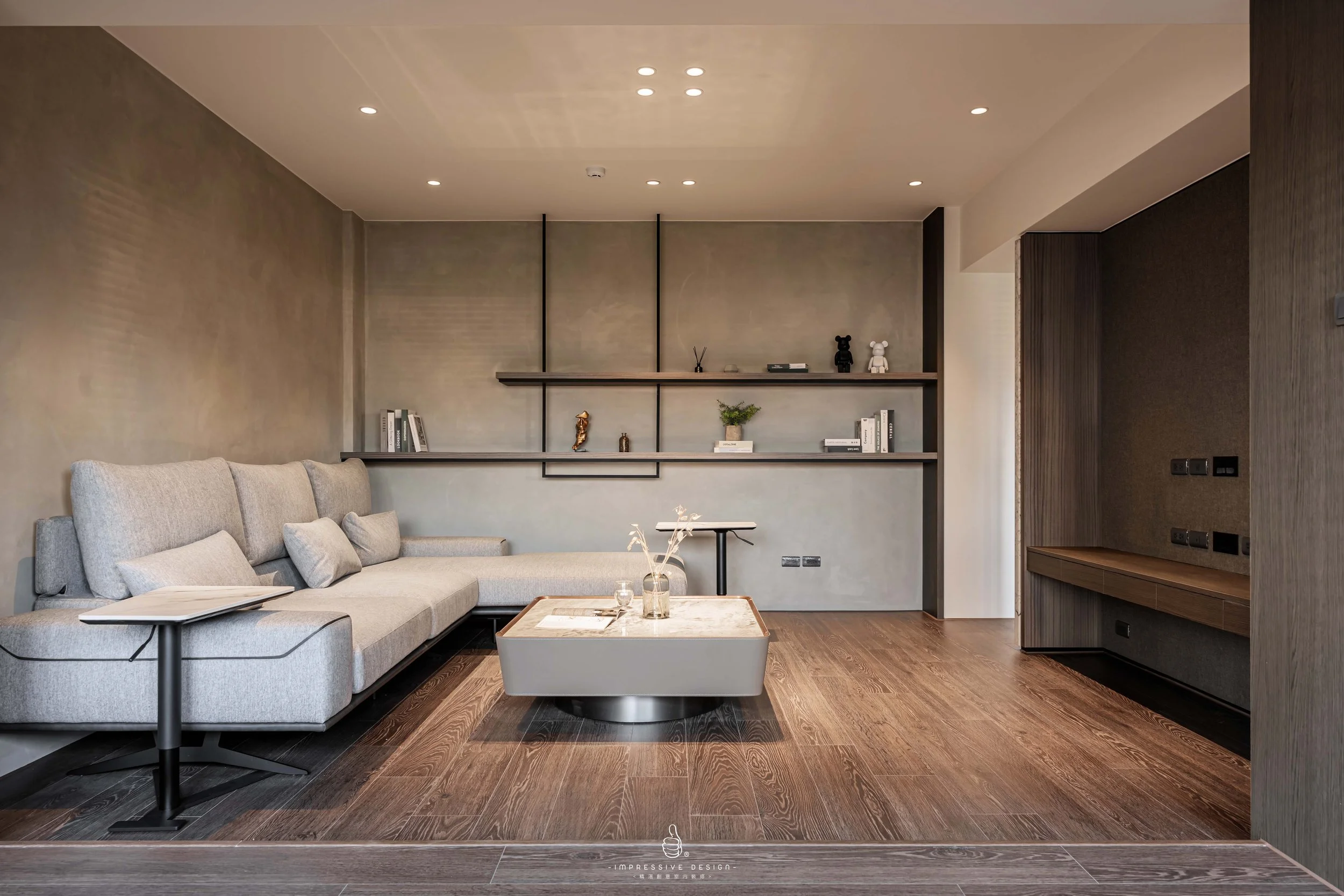 Modern living room with a light gray sofa, wooden coffee table, and built-in wall shelving with decorative objects. The room features dark wood flooring, recessed ceiling lights, and a neutral wall color.