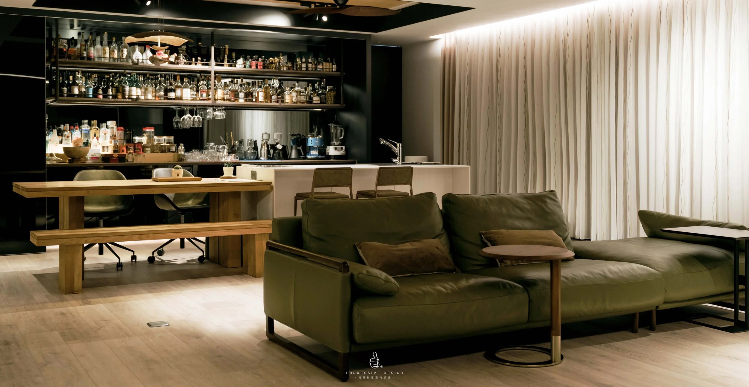 Living room with a green sofa, wooden side table, and large window with curtains; background features a bar area with shelves of alcohol bottles and glassware, and a kitchen counter with chairs.