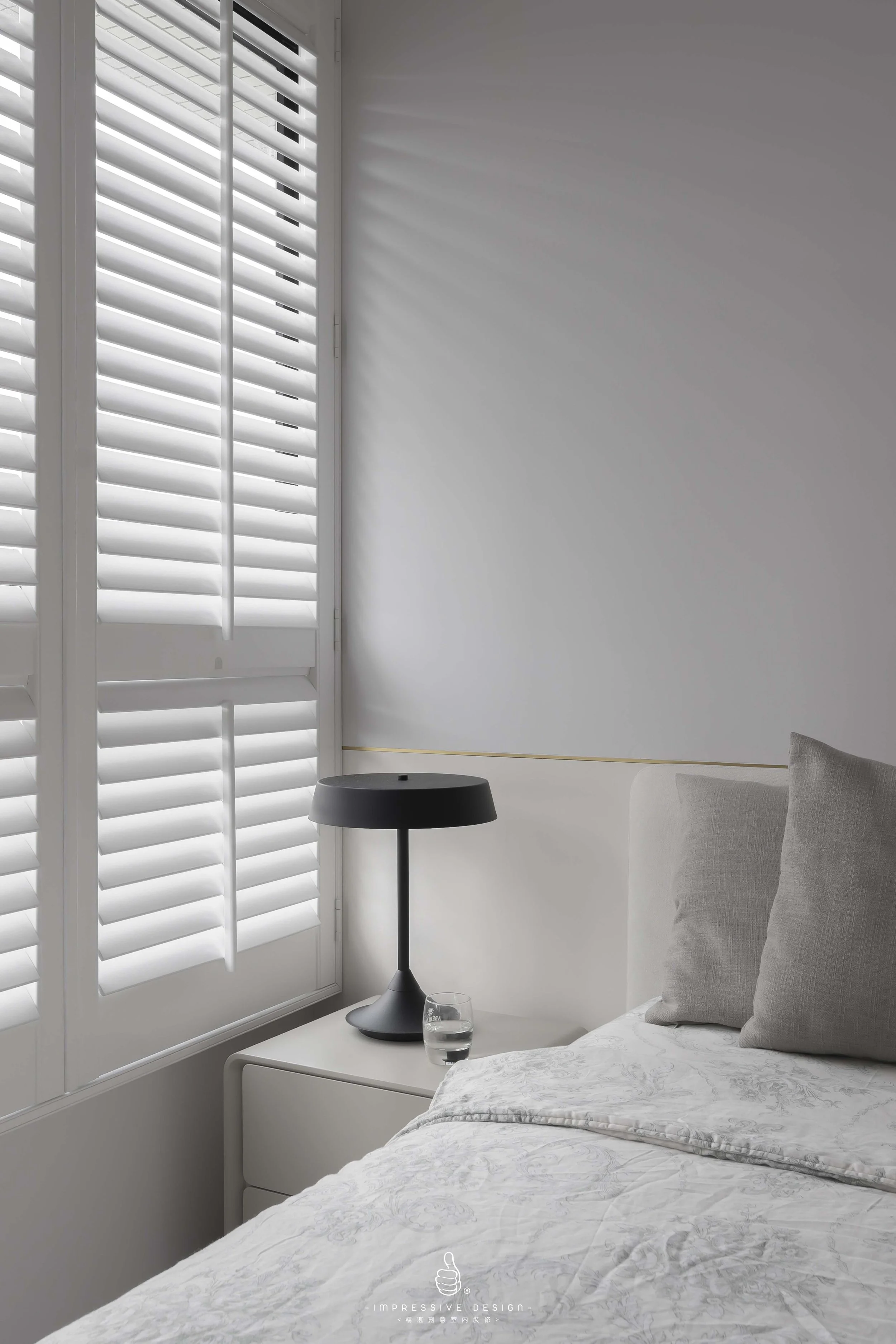 A bedroom corner with a white bed, beige pillows, a black bedside lamp, a glass of water, and a window with white shutters casting shadows on the wall.