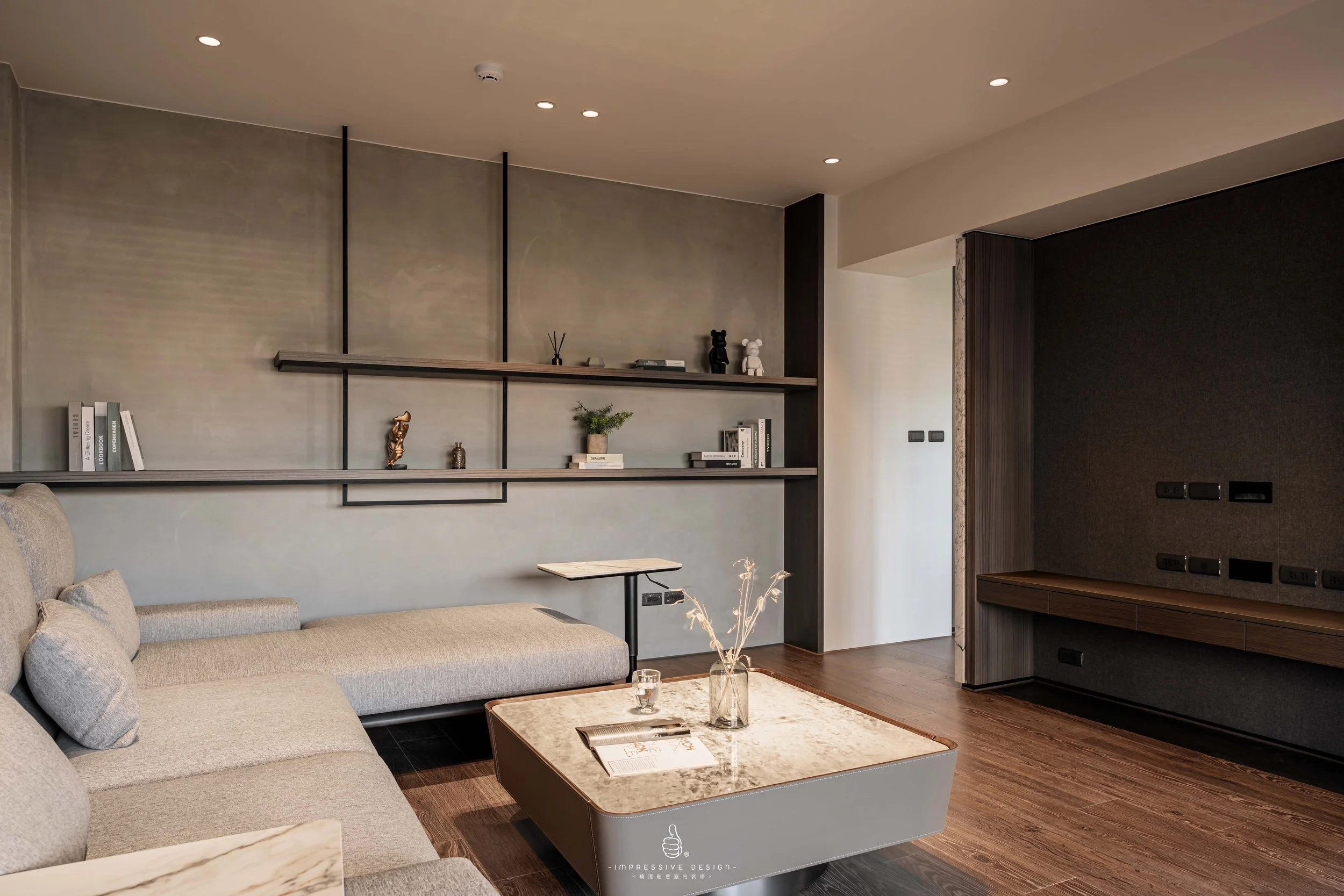 Modern living room with gray sofa, marble coffee table, wooden shelves with decor, and a dark feature wall with outlets.