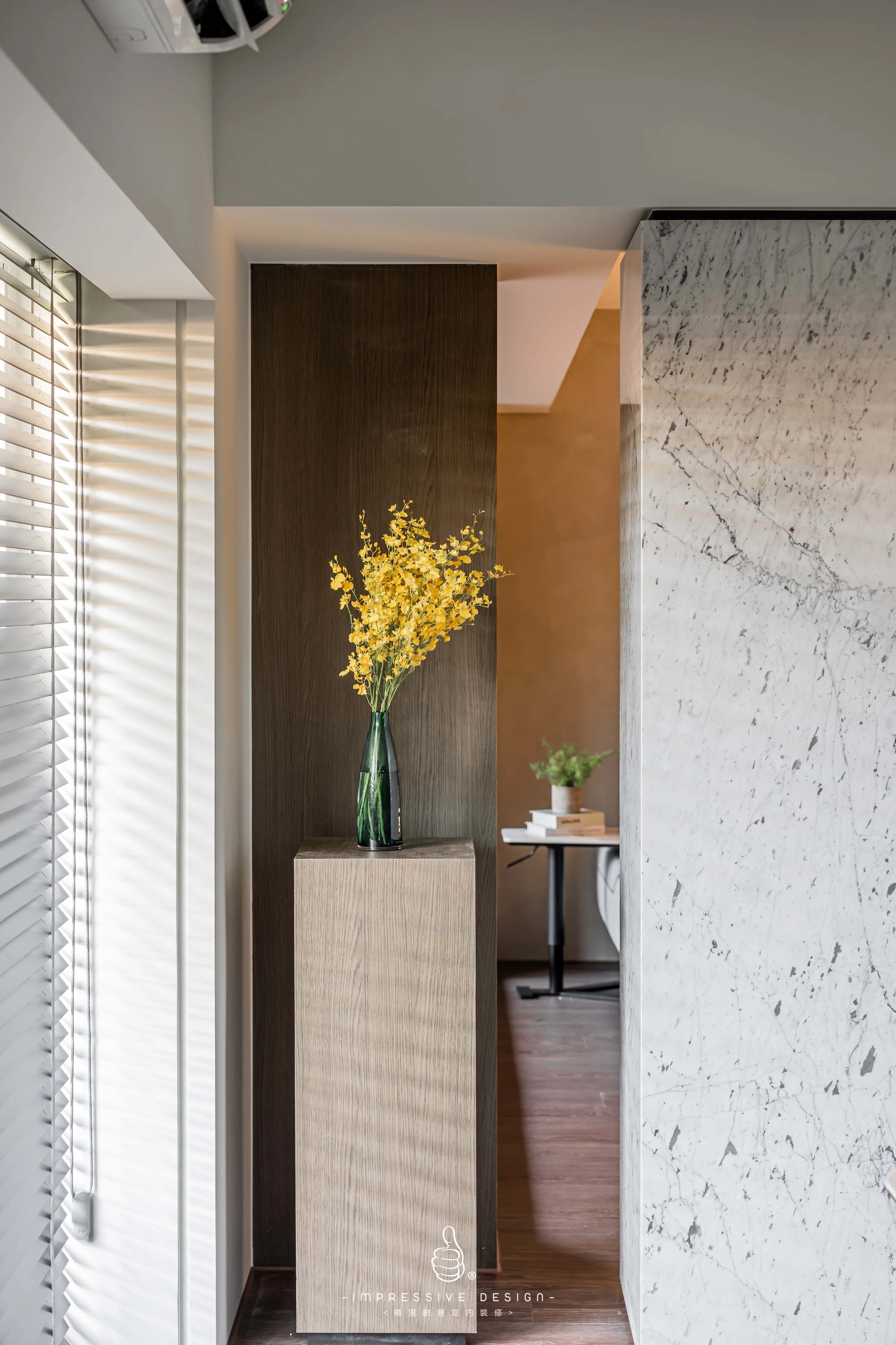 A modern interior space with a tall green vase holding yellow flowers on a wooden pedestal against a dark wood panel, with a light-colored marble wall on the right and a window with horizontal blinds on the left. In the background, a small table with