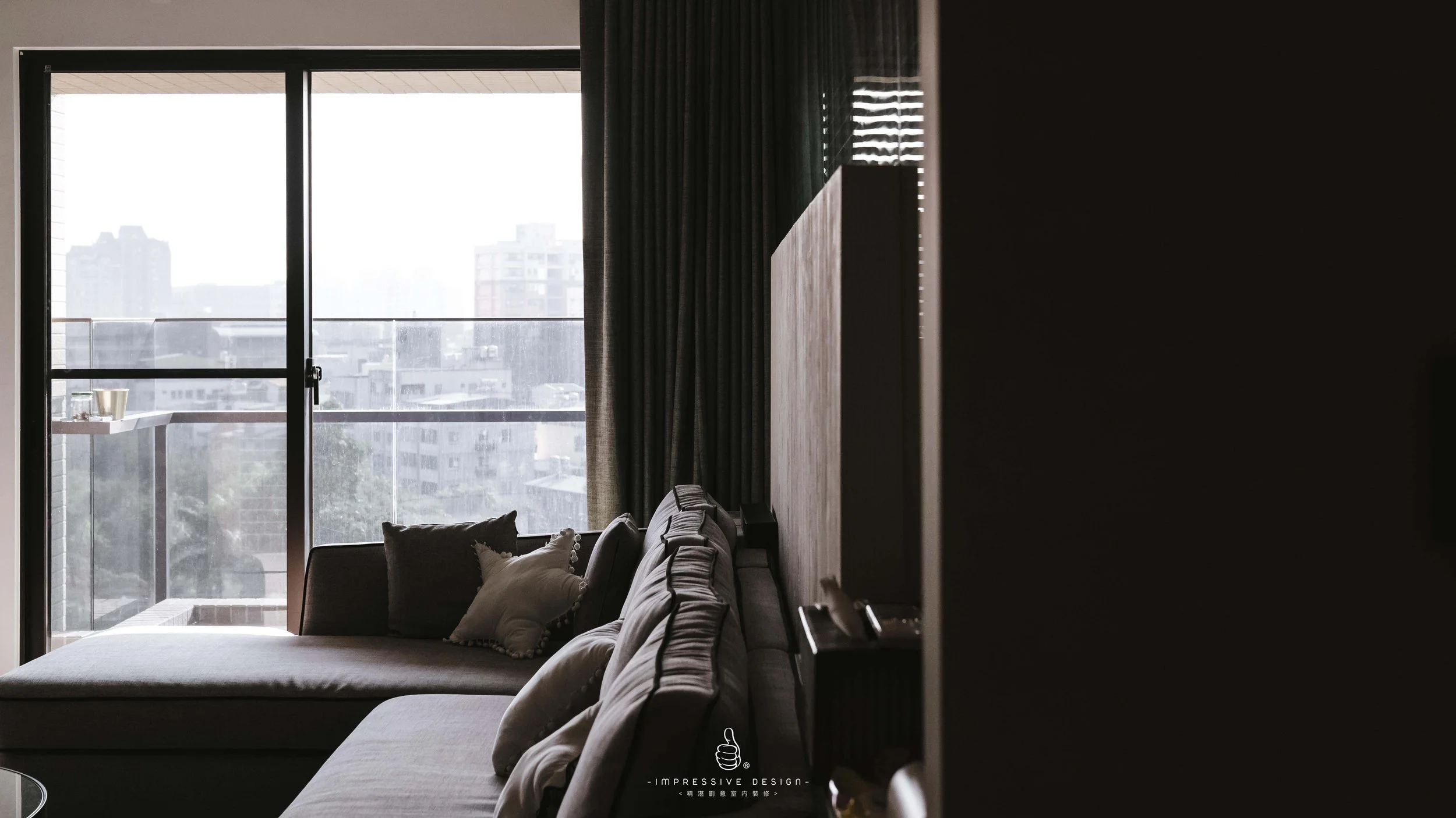 Living room with a sofa and pillows, large sliding glass door leading to a balcony, tall city buildings outside, curtains partially covering the window.