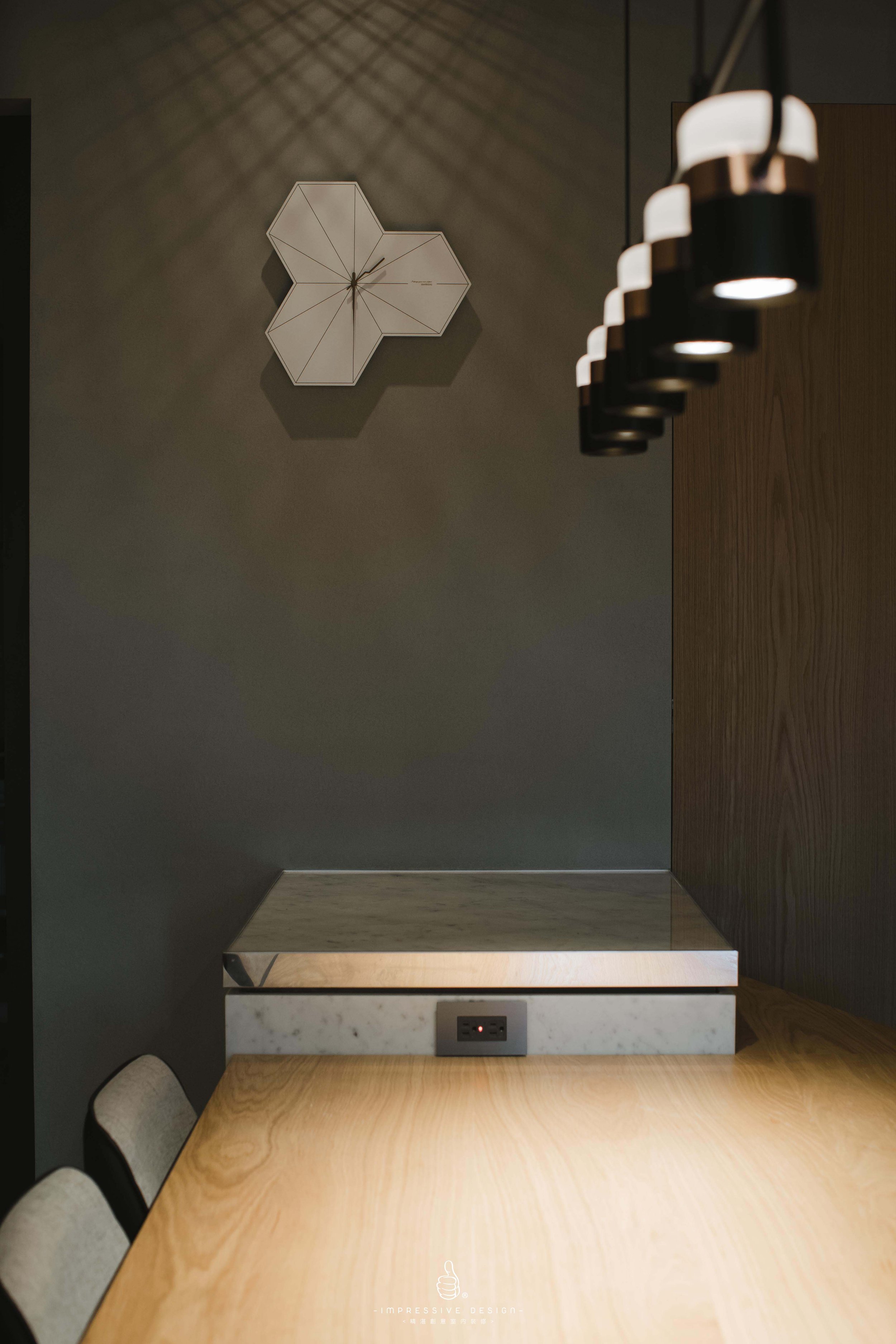 Interior view of a modern dining or conference area with a wooden table, beige upholstered chairs, a dark accent wall, a decorative wall clock, and a line of hanging pendant lights.