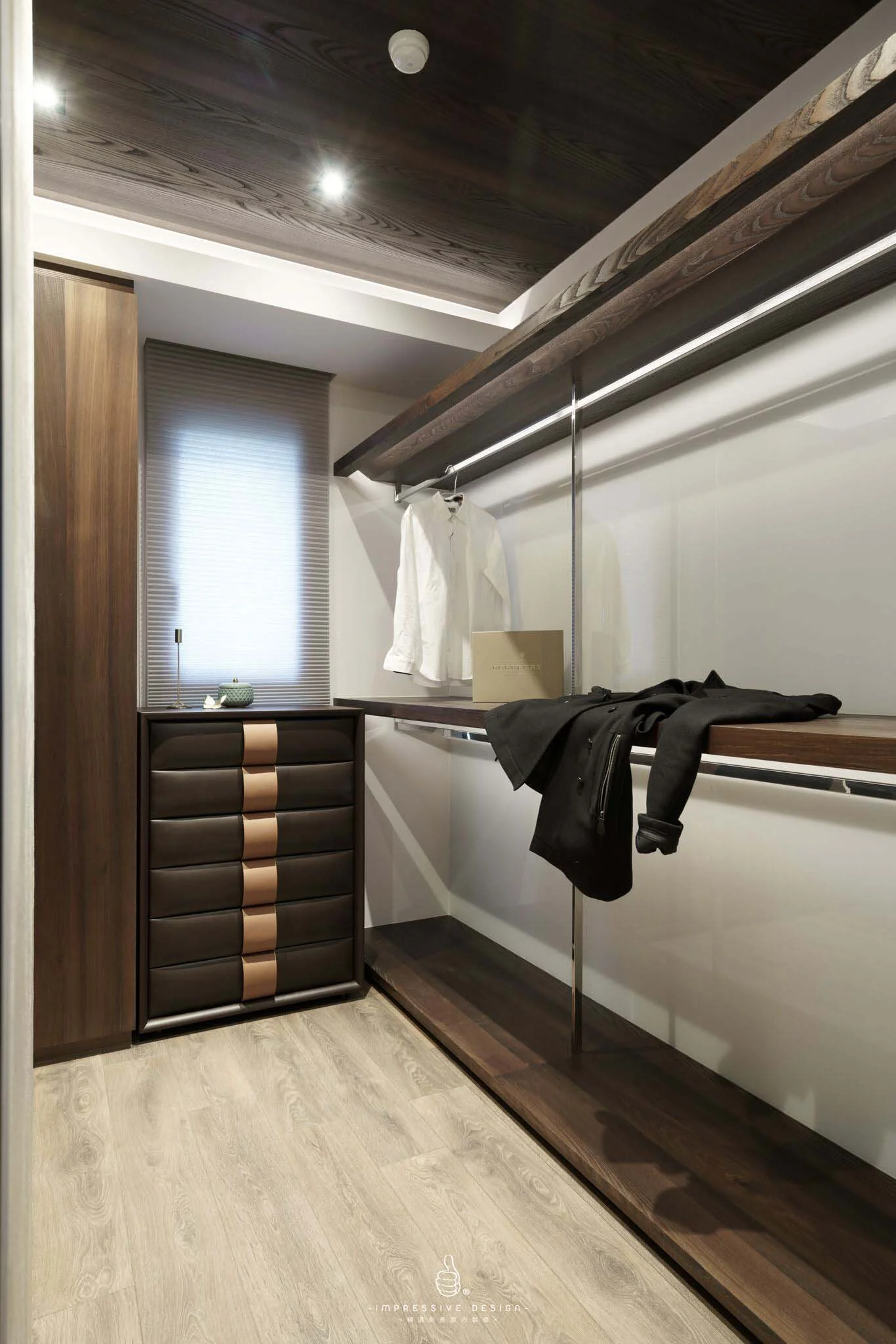 A modern walk-in closet with wooden storage units, a small chest with drawers, hanging clothes, and a window with blinds.