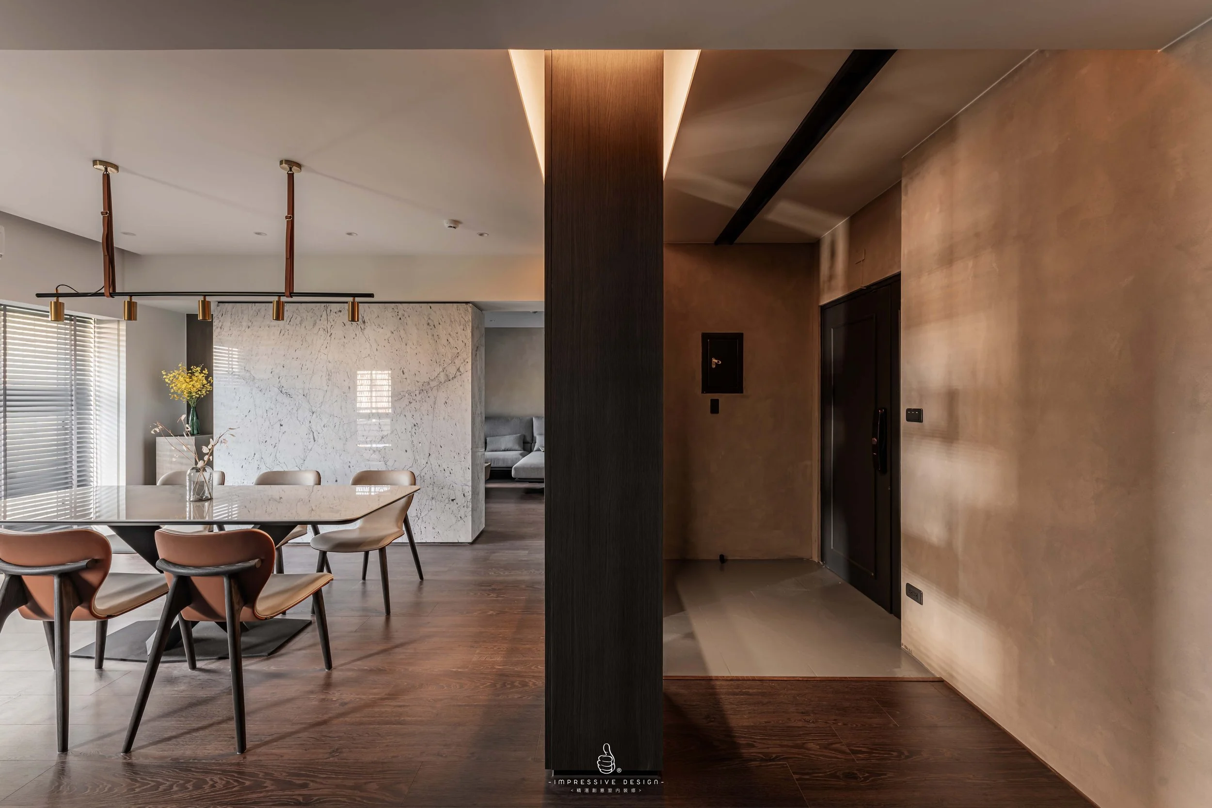 Interior of a modern home with a dining area and living room, split by a dark wooden column, featuring wooden floors, a large marble wall, and minimalist decor.