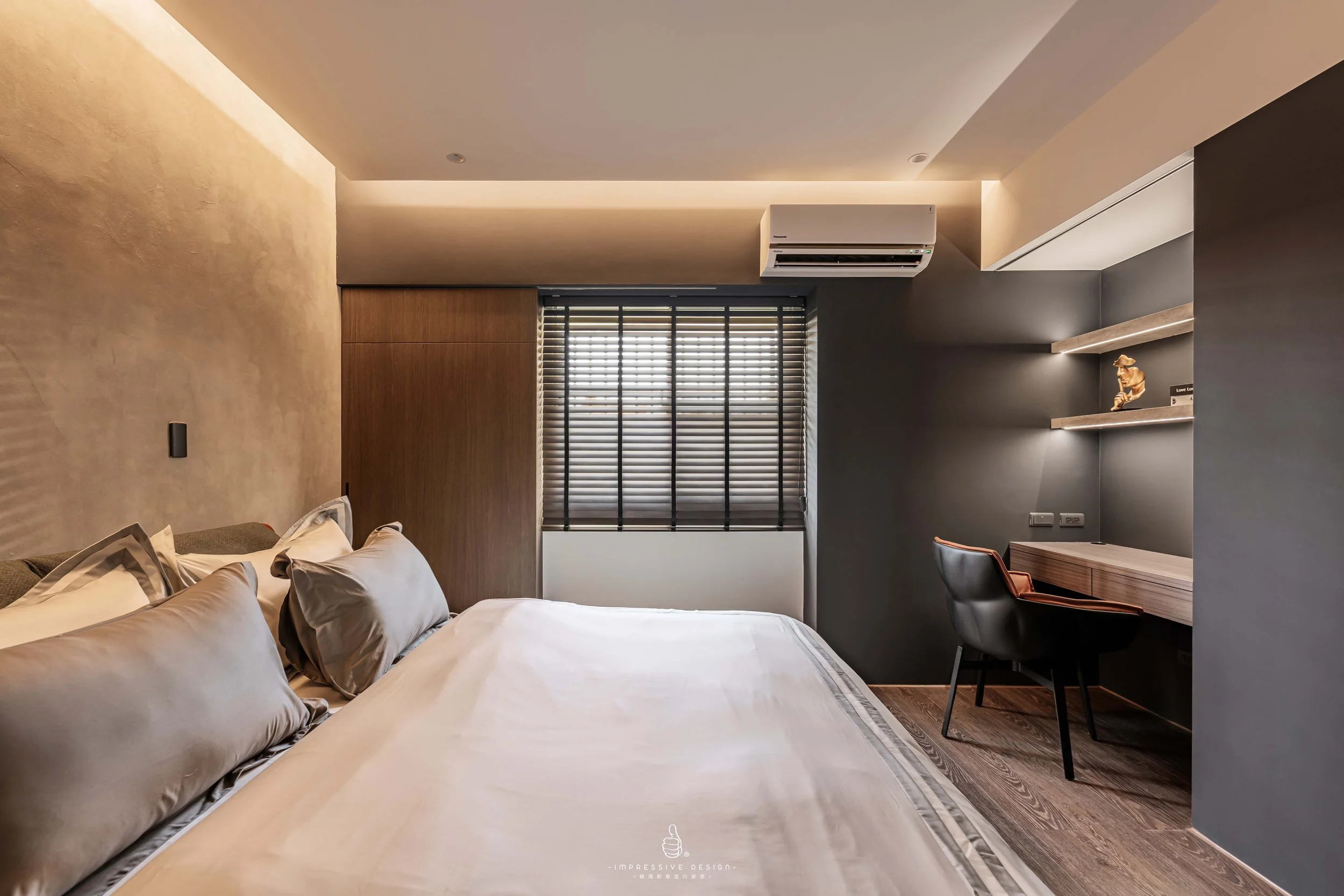 Modern bedroom with a large bed, gray and beige bedding, wooden accents, a window with blinds, a wall-mounted air conditioner, a small desk with a chair, and decorative shelves.