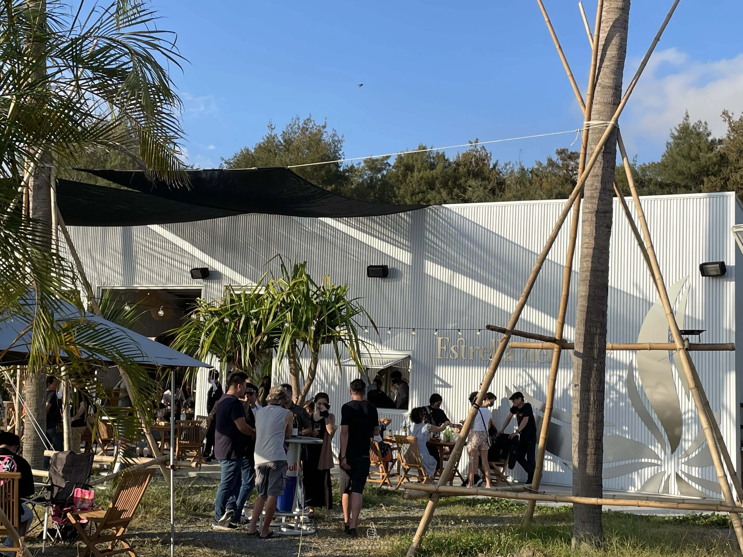 Outdoor scene with people gathered around tables at a restaurant or cafe, with palm trees and a white building in the background. There are string lights, a black sunshade, and a large wooden teepee-like structure near a palm tree.