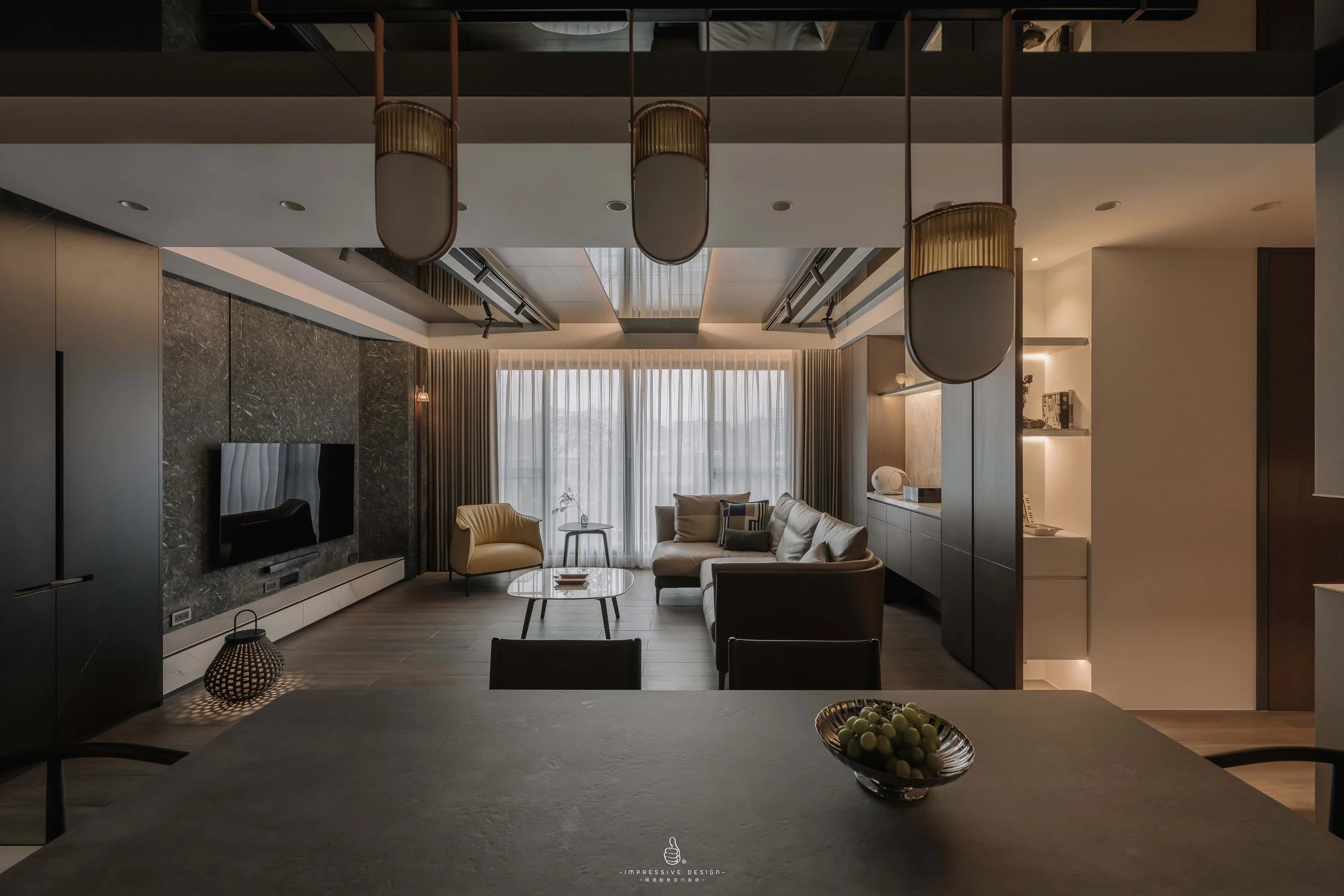 Modern living room with a sofa, armchair, coffee table, and TV, illuminated by natural light from large window with curtains, and featuring ceiling lights and decorative shelving.