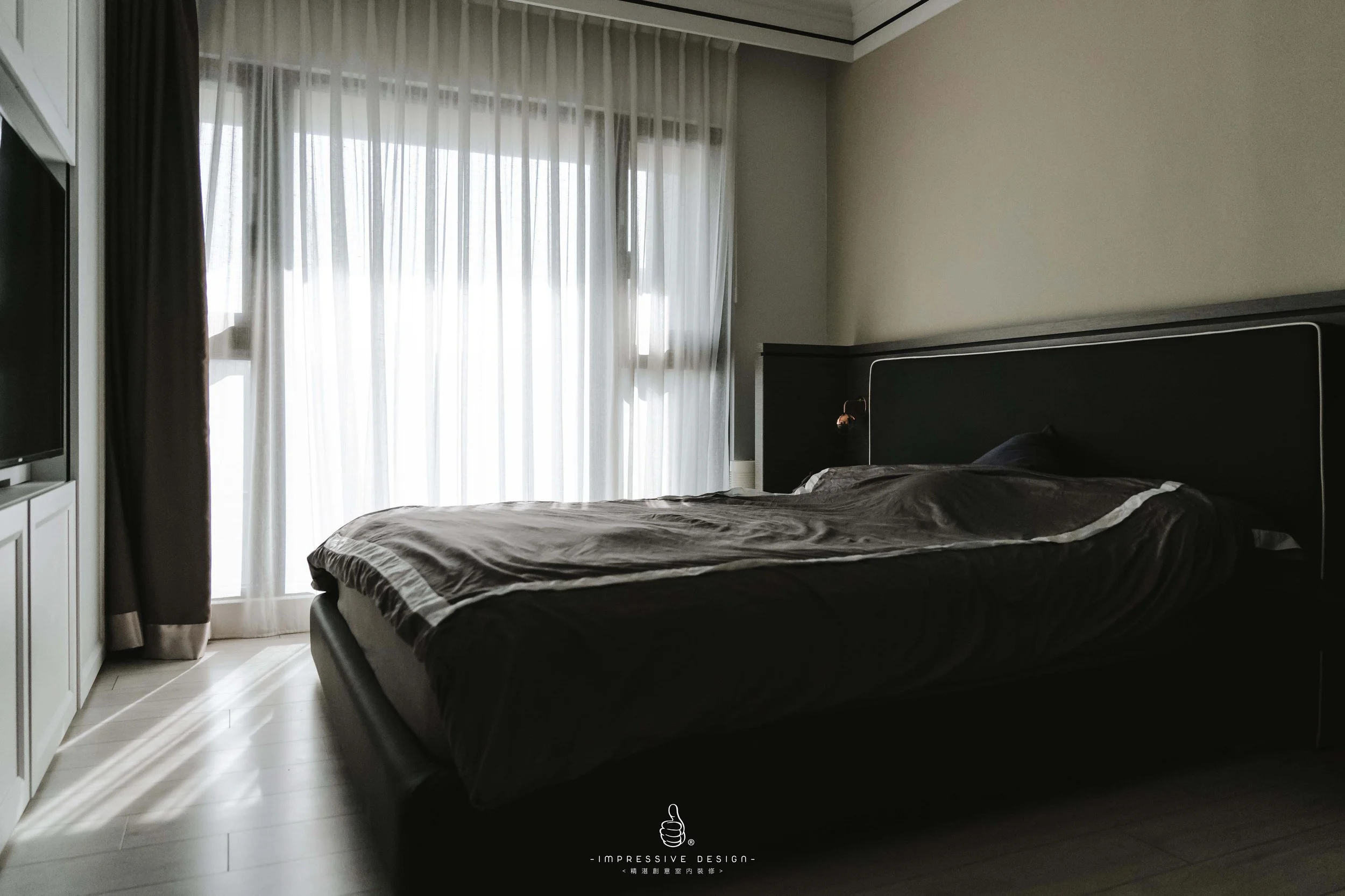 A bedroom with a bed covered in dark gray bedding and a black headboard. Light streams in through sheer white curtains covering large windows, with a darker curtain pulled to the side. A television is partially visible on the left side of the room.