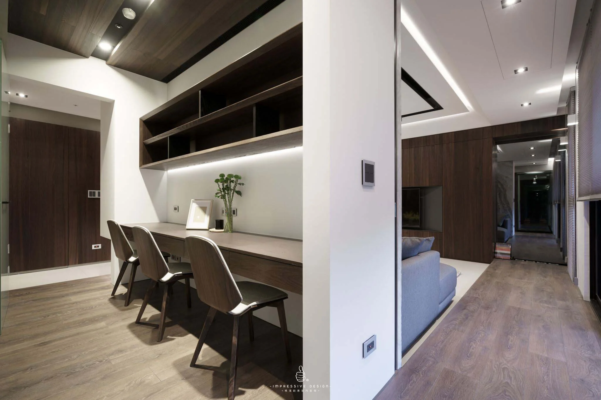 Split view of modern living space with a workspace featuring a desk, chairs, and wall shelves on the left, and a cozy sitting area with a sofa and TV on the right.