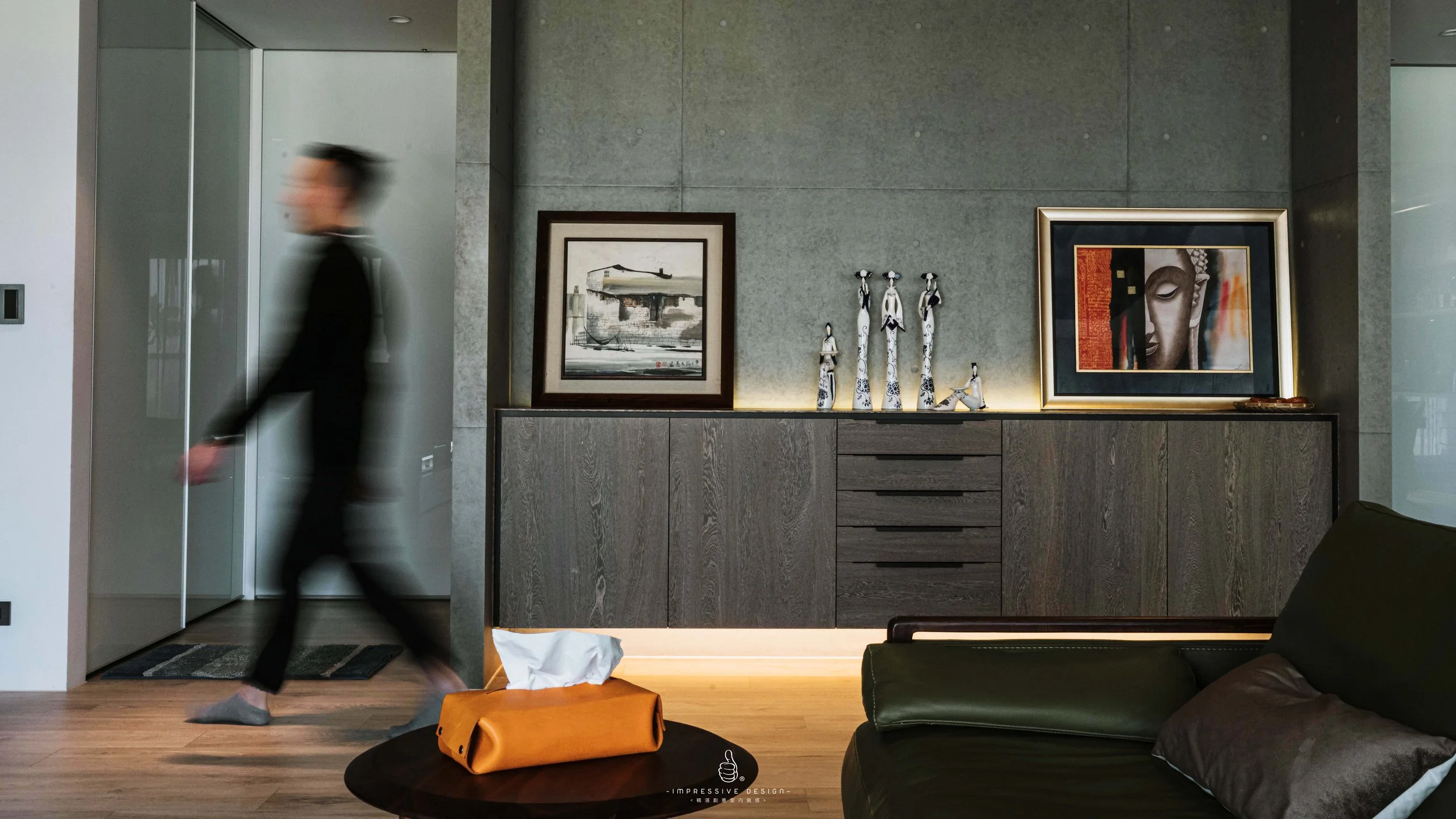 A blurred person in black attire walking across a modern living room. On the wall behind them are framed artwork and decorative figures on a wooden cabinet. There is a green sofa with pillows and a small table with a tissue box in the foreground.