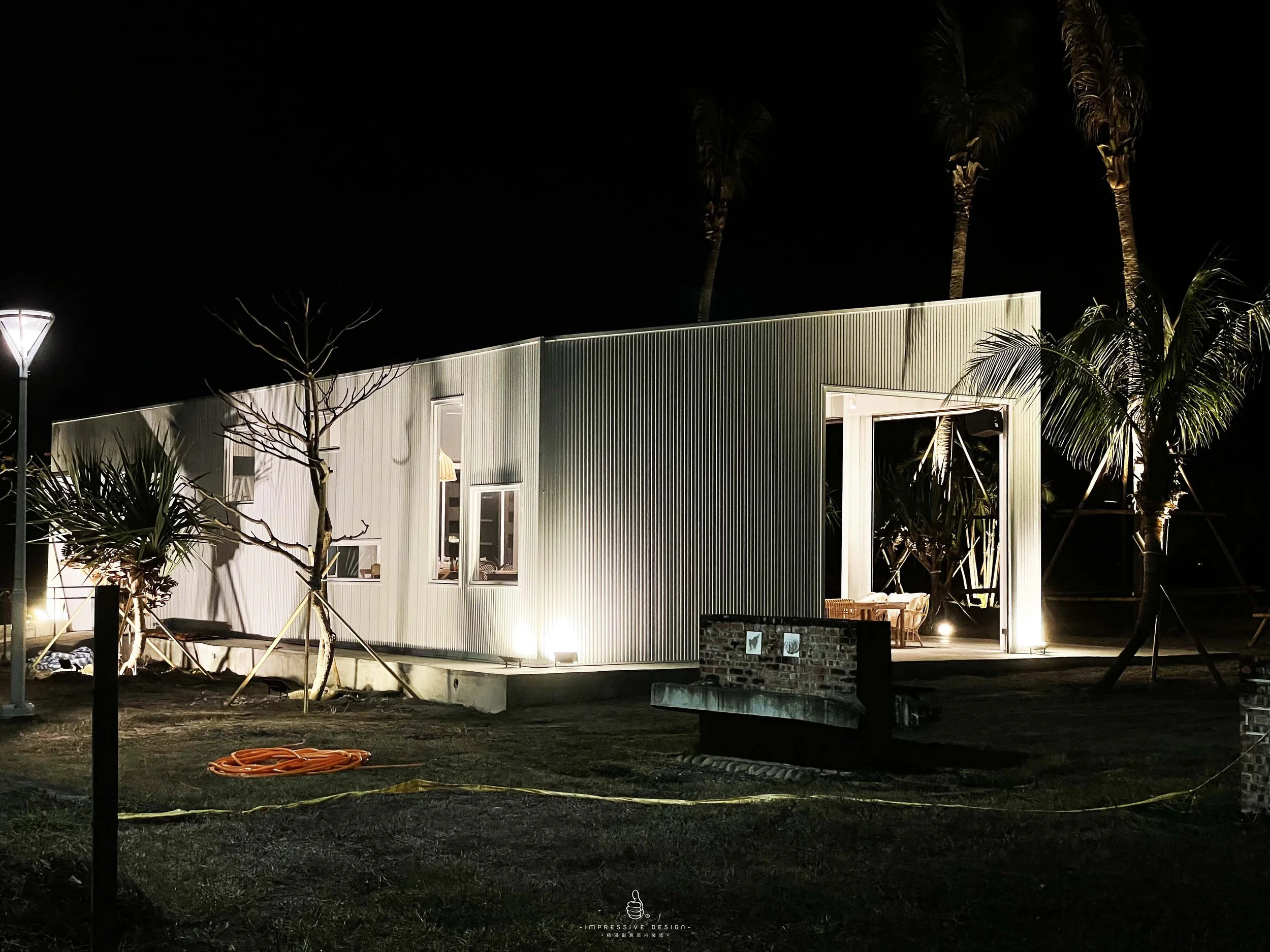 Modern house at night with exterior lighting, surrounded by palm trees and a leafless tree, a streetlamp on the left, orange hose and yellow cable on the ground, windows and an open door visible.
