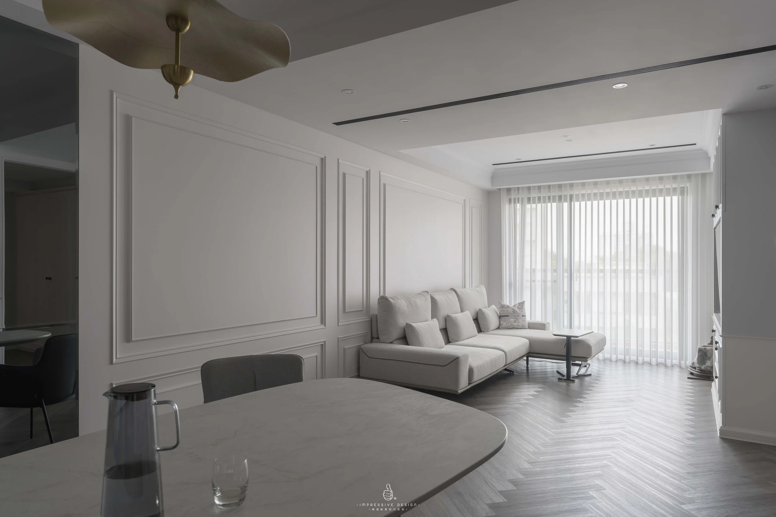 Bright living room with white walls, a light-colored sectional sofa with multiple cushions, large windows with sheer curtains, dark wood herringbone floor, and part of a marble table with a pitcher and glass in the foreground.