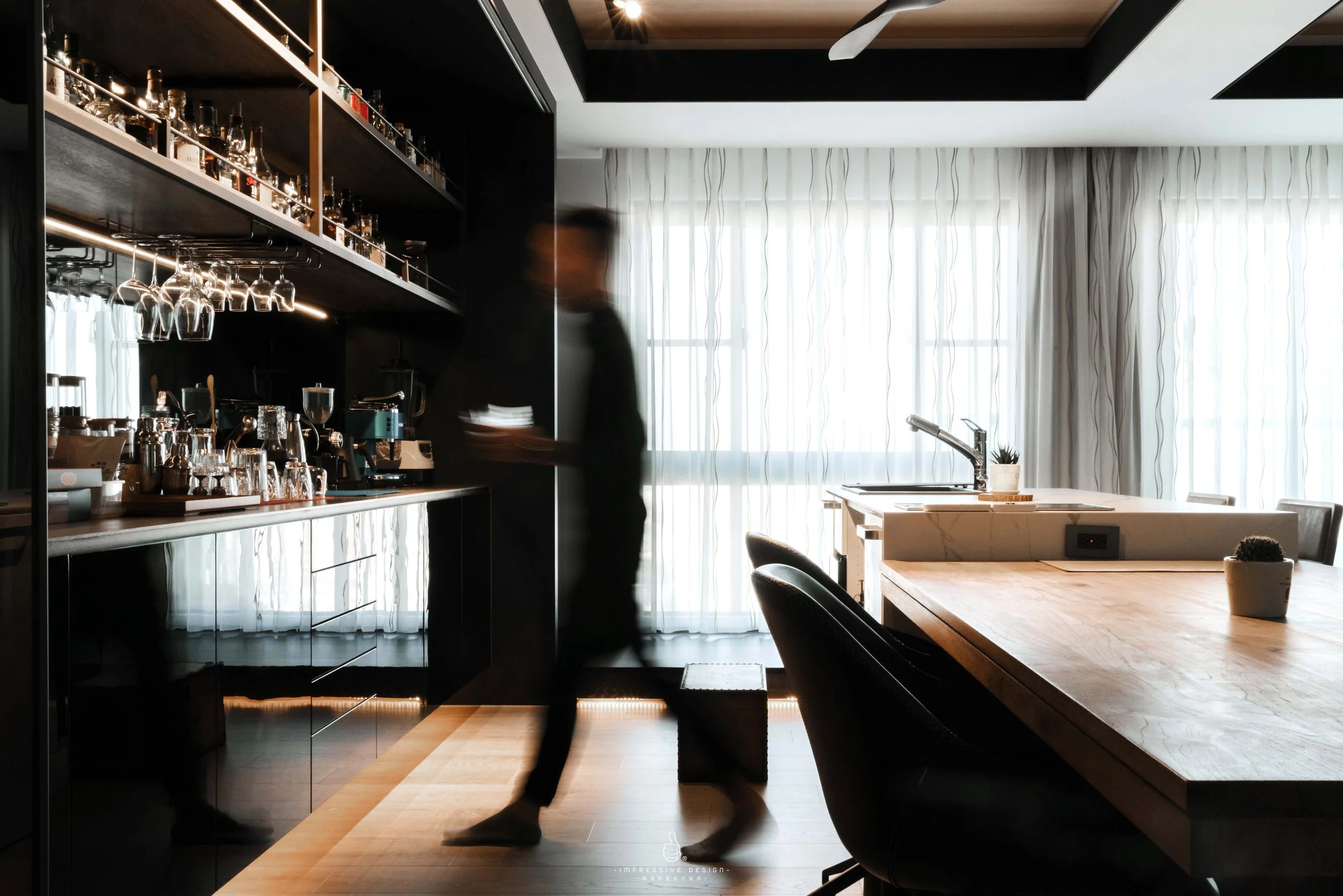 A person stands near a bar counter in a modern kitchen with large windows and sheer curtains. The kitchen has a wooden dining table, black chairs, and various glassware and bottles on open shelves. The room is illuminated by natural light.