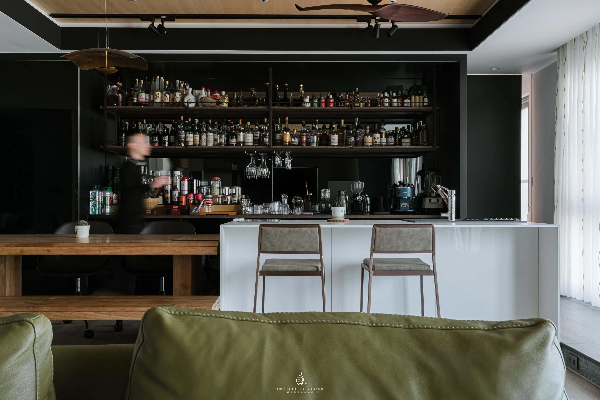 Modern kitchen with a white island counter, two bar stools, a black and wooden shelving unit with bottles and glasses, and a wooden dining table with a small plant. A blurred person is behind the counter, and a large window with white curtains is on 