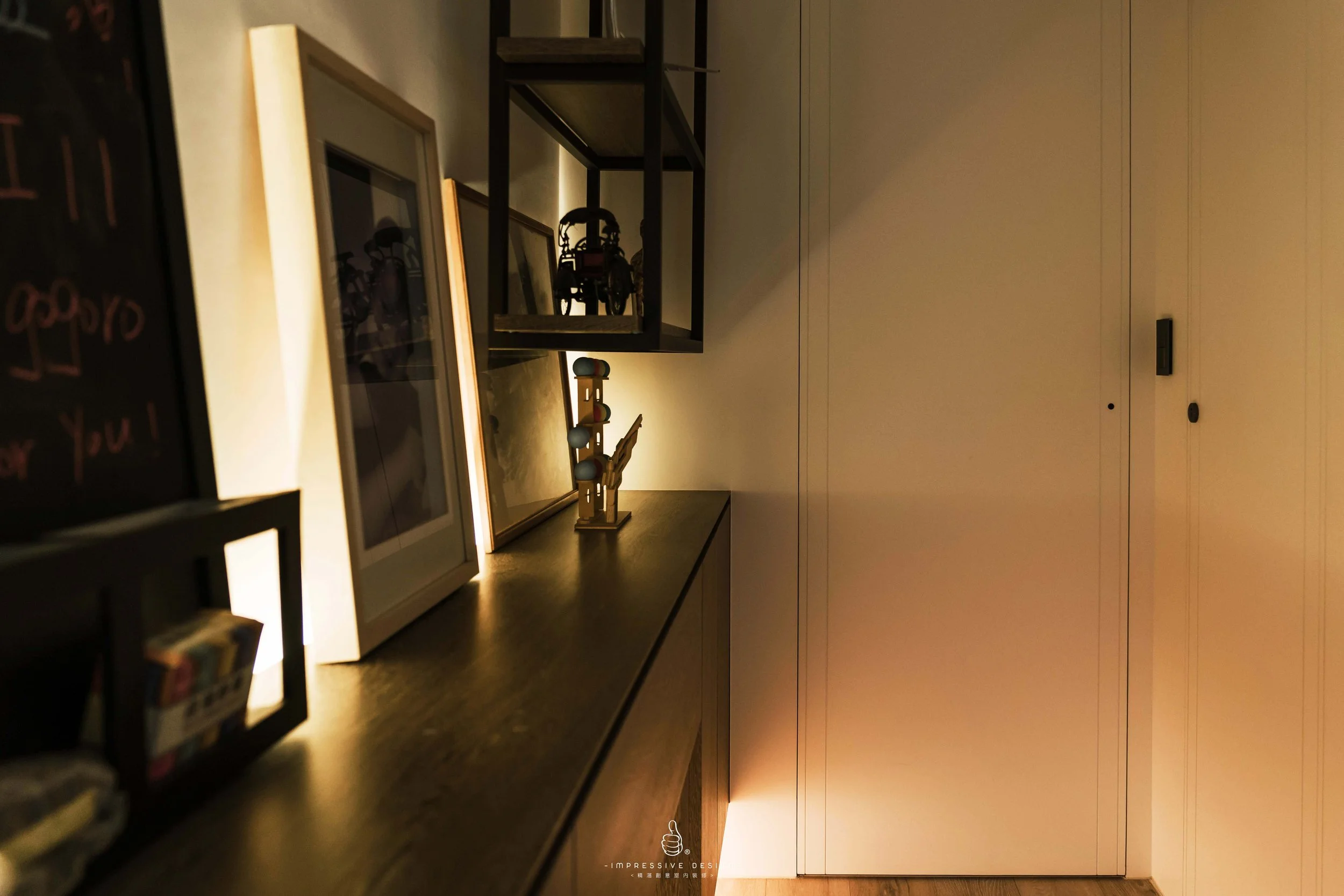 A lit display shelf with framed photos and small decorative objects on a dark wooden surface, against a white wall with built-in cabinets.
