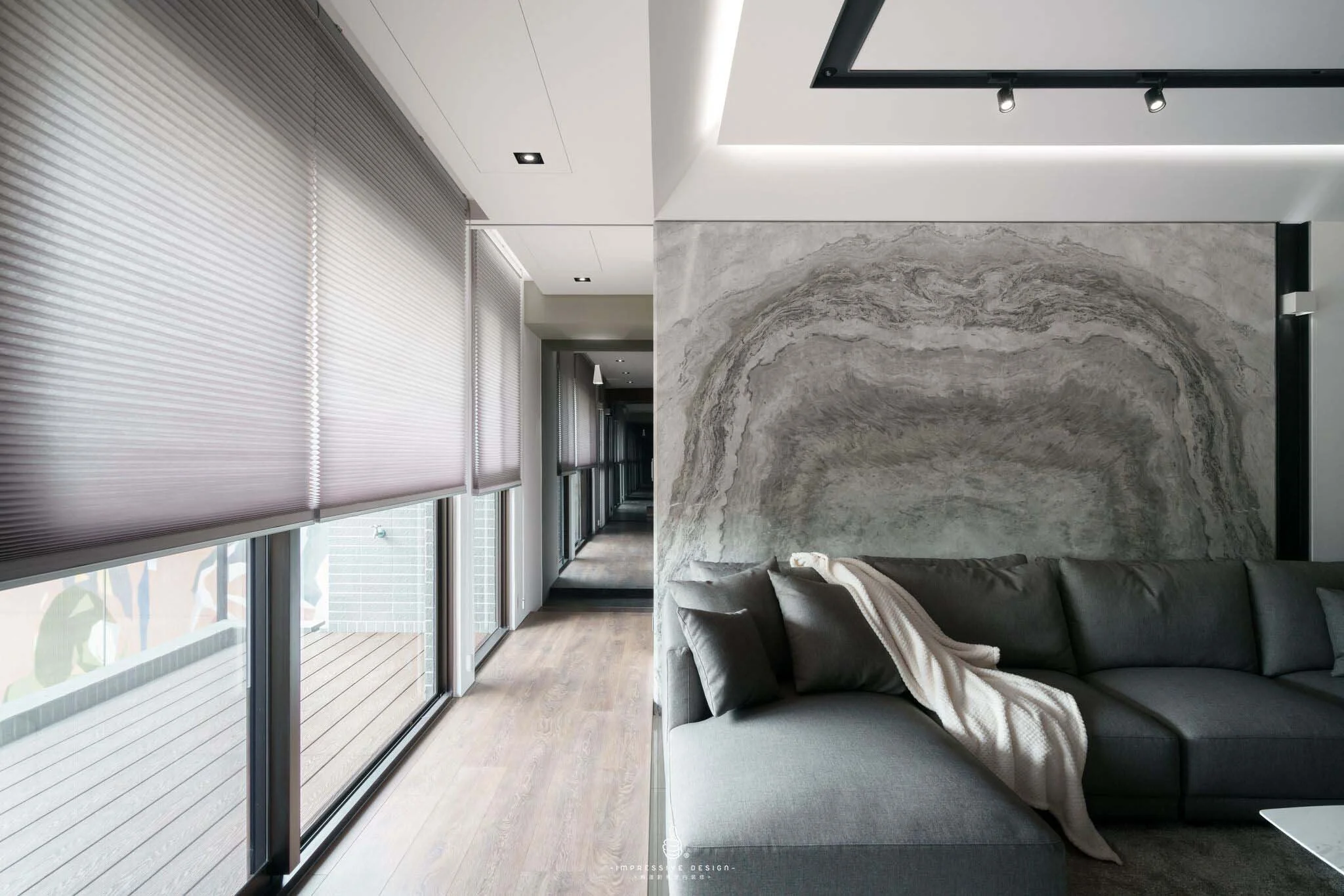 Modern living room with large windows, gray roller blinds, a gray sectional sofa with a blanket, and a textured gray wall.