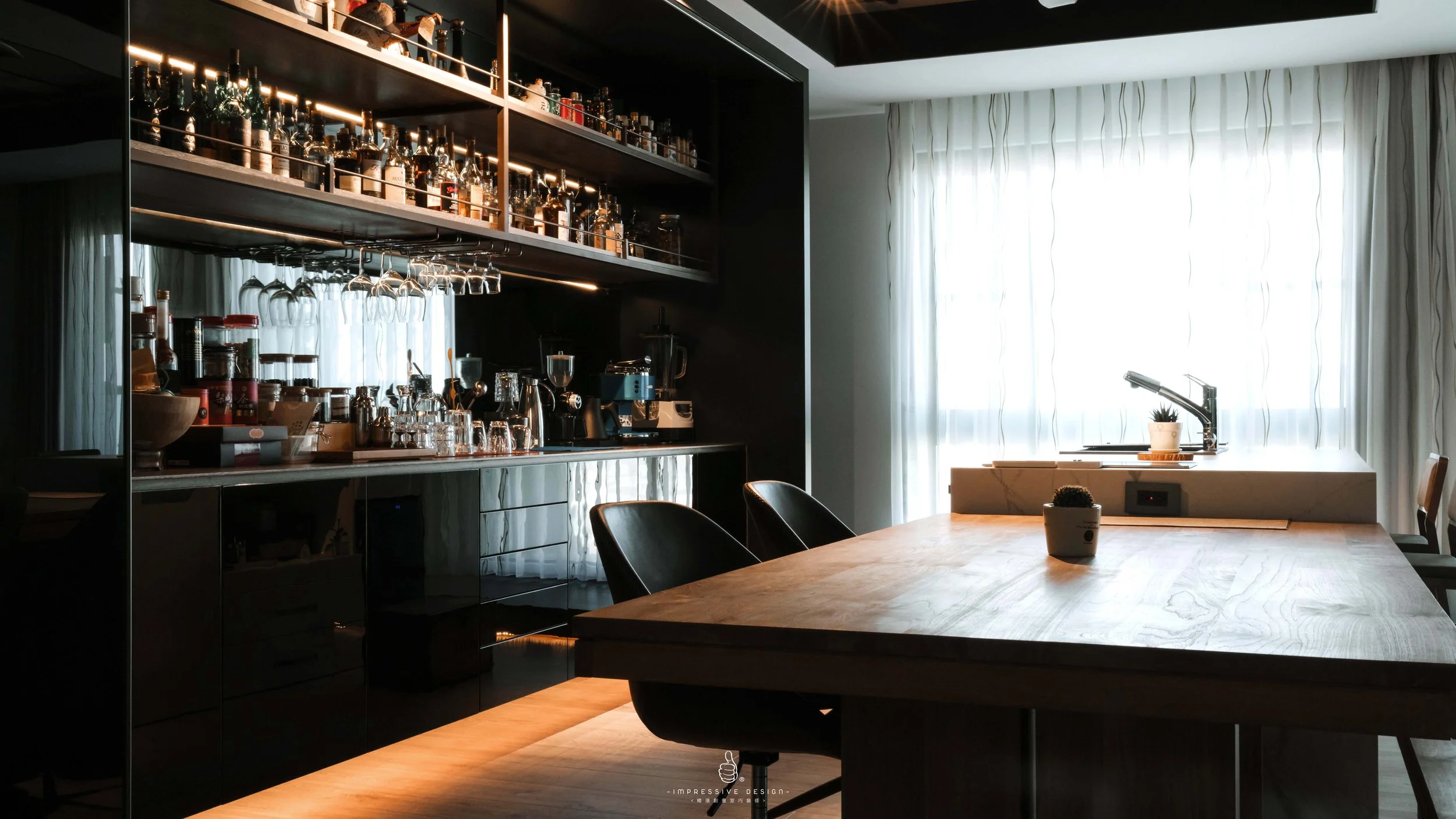 Modern kitchen with a large wooden dining table, black chairs, and a bar area with glasses and bottles. Bright natural light coming through sheer curtains on a window, with a small potted plant on the table.