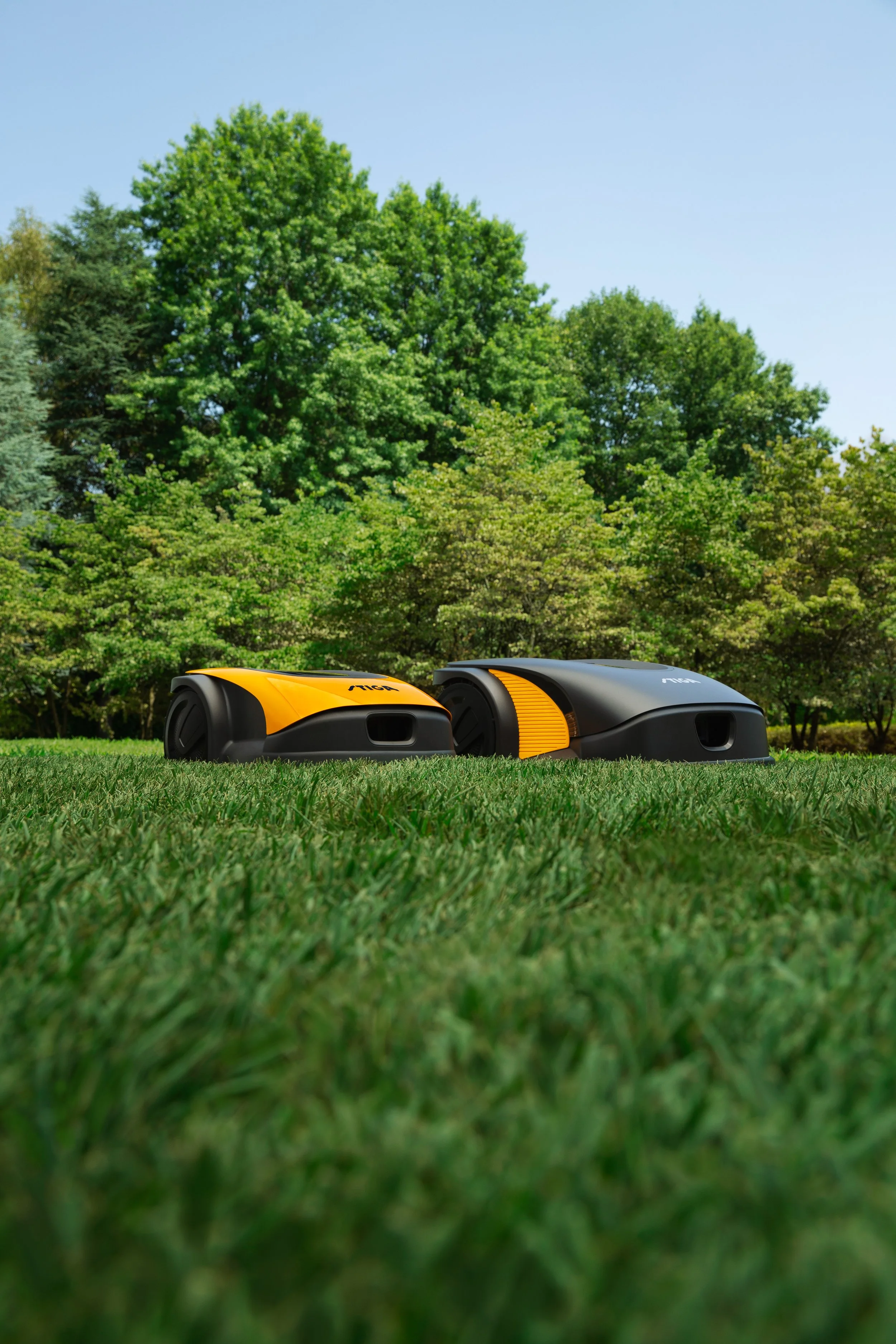 Two robotic lawn mowers on a well-maintained grassy lawn with trees and blue sky in the background.