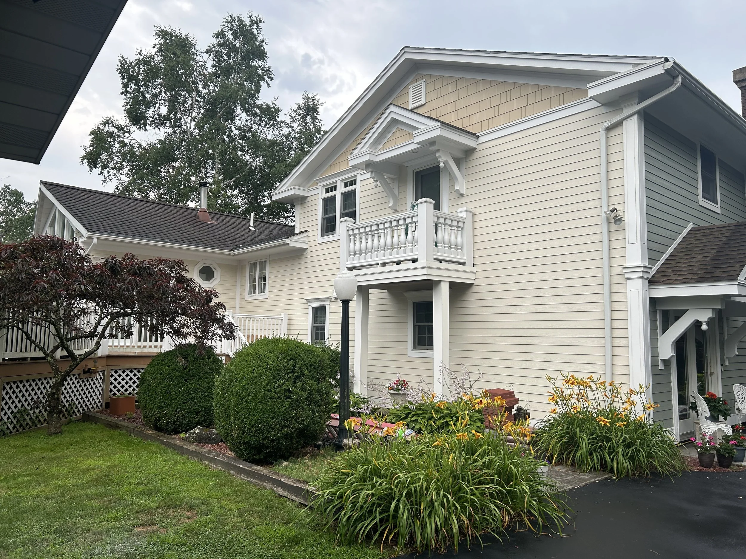A two-story house with beige siding and white trim, surrounded by a landscaped yard with bushes, flowers, and a tree, under an overcast sky.