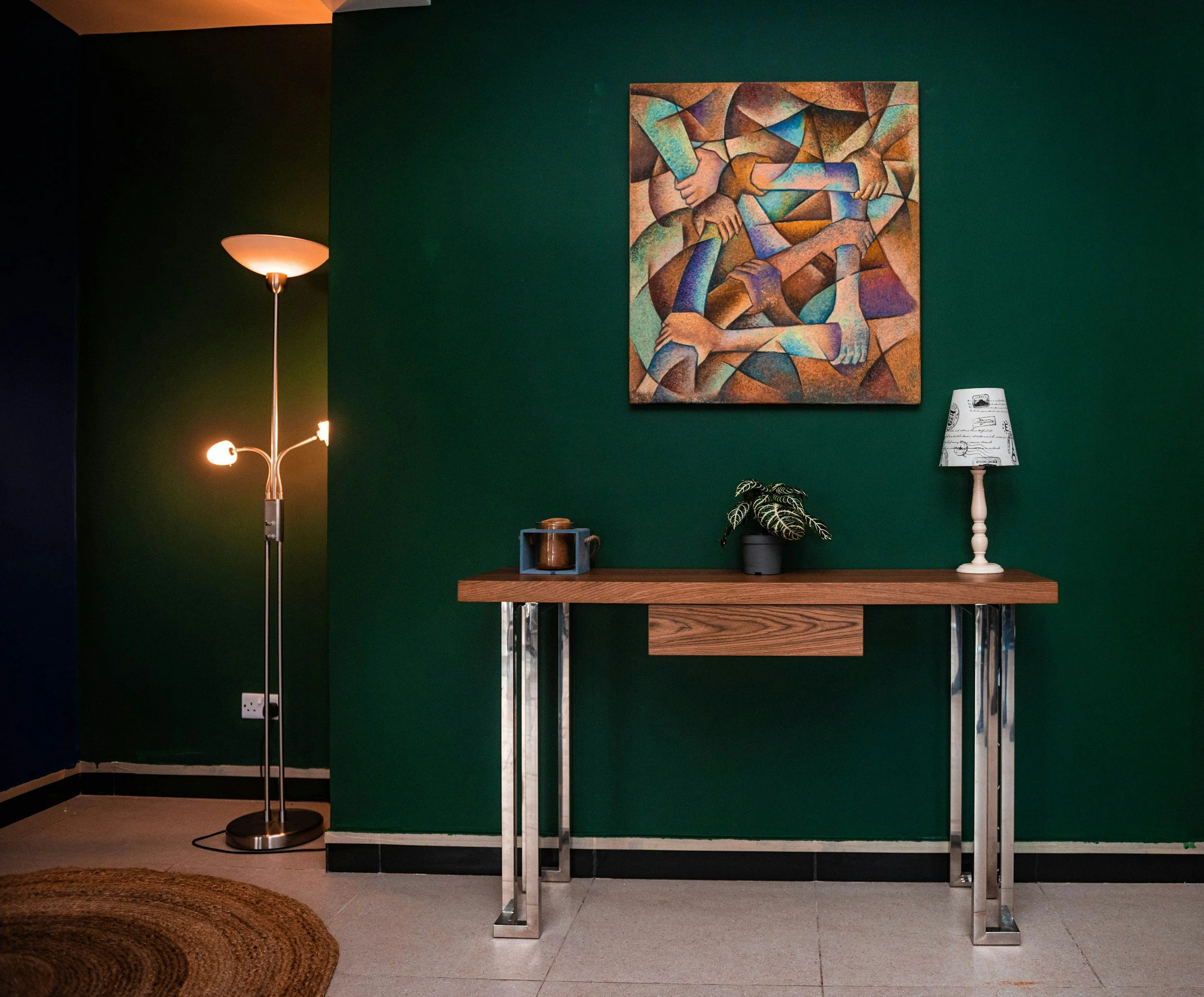 A room with dark green walls, a painting depicting multi-colored abstract figures, a white desk lamp with a poster print lampshade, a small table with a potted plant, a wooden table, and a silver floor lamp.