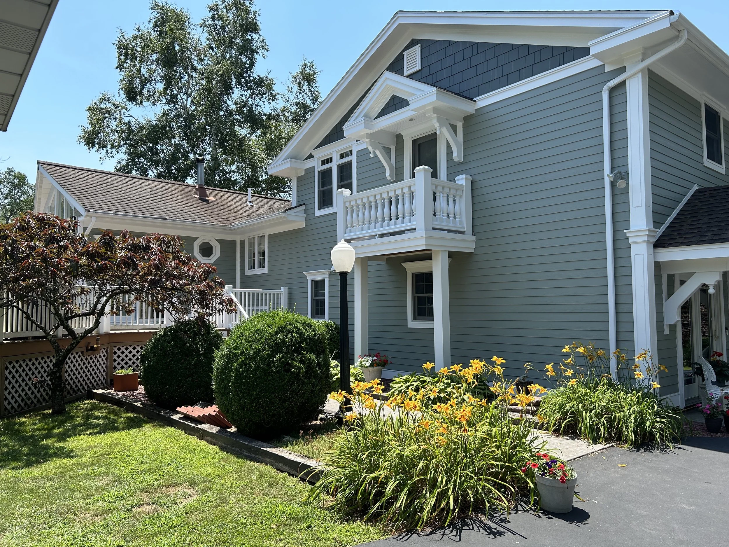 Side view of a blue multi-story house with white trim, surrounded by a well-maintained garden with blooming flowers and a small tree, under a clear blue sky.