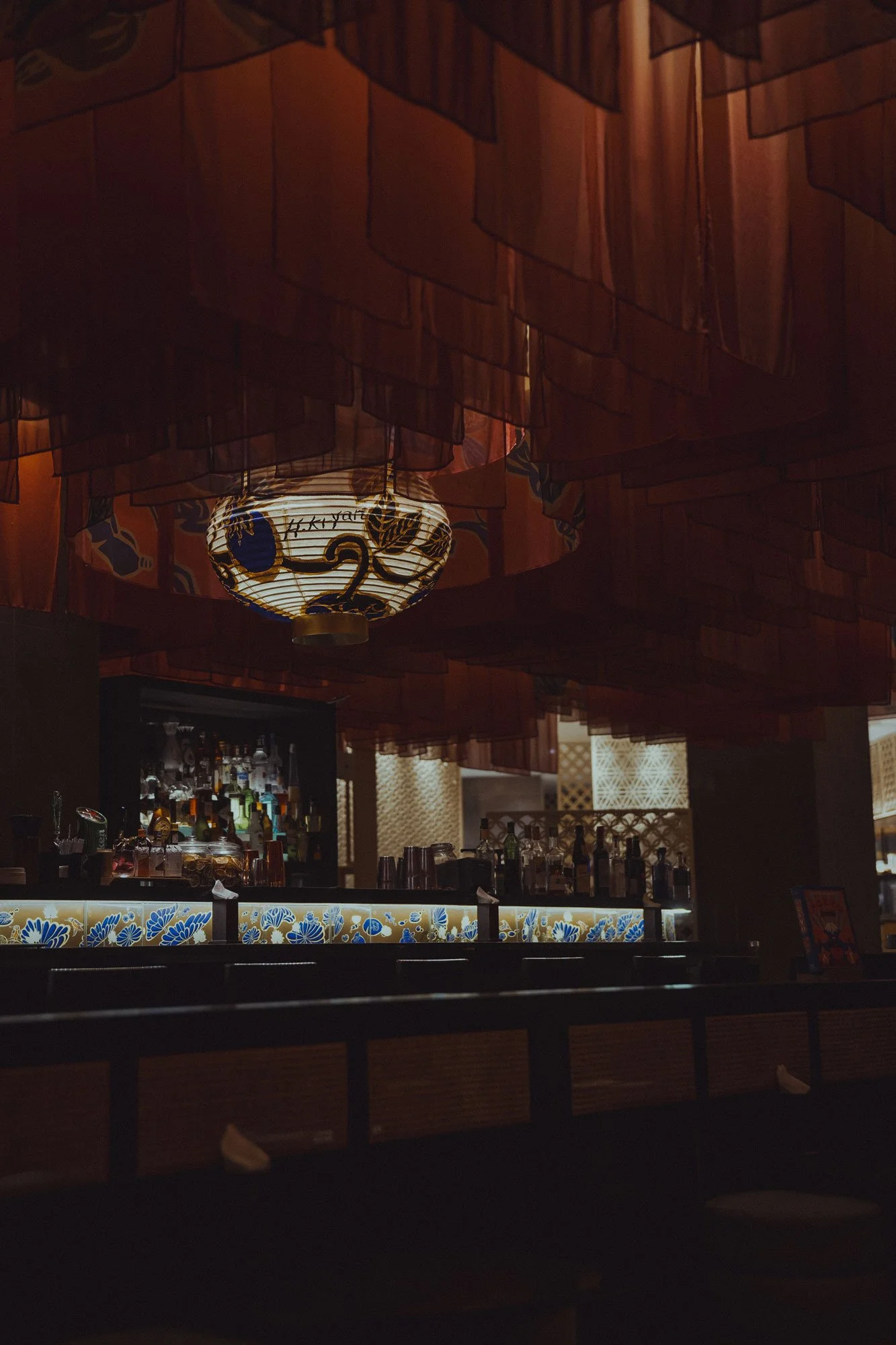 Dimly lit bar area with hanging fabric or paper decorations on the ceiling, a central lantern-style light fixture, and bottles of alcohol on the shelves behind the bar counter.