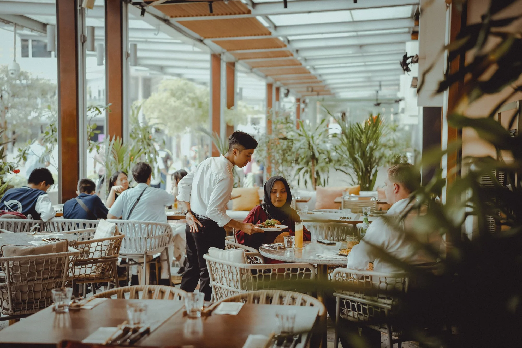 A busy restaurant with patrons dining and staff serving. Several people are seated at tables, and a waitress is serving a dish to a woman in a hijab. The restaurant has large windows, indoor plants, and a modern decor.