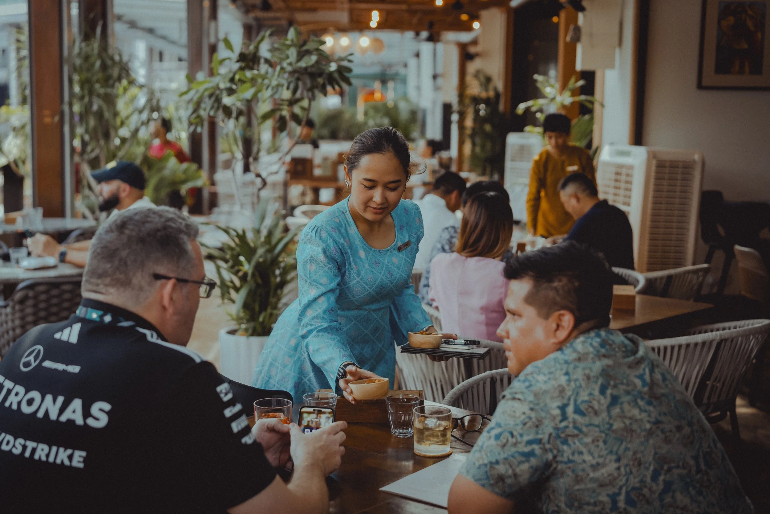 A waitress serving food to customers at a restaurant. The restaurant has large windows, potted plants, and several patrons seated at tables.