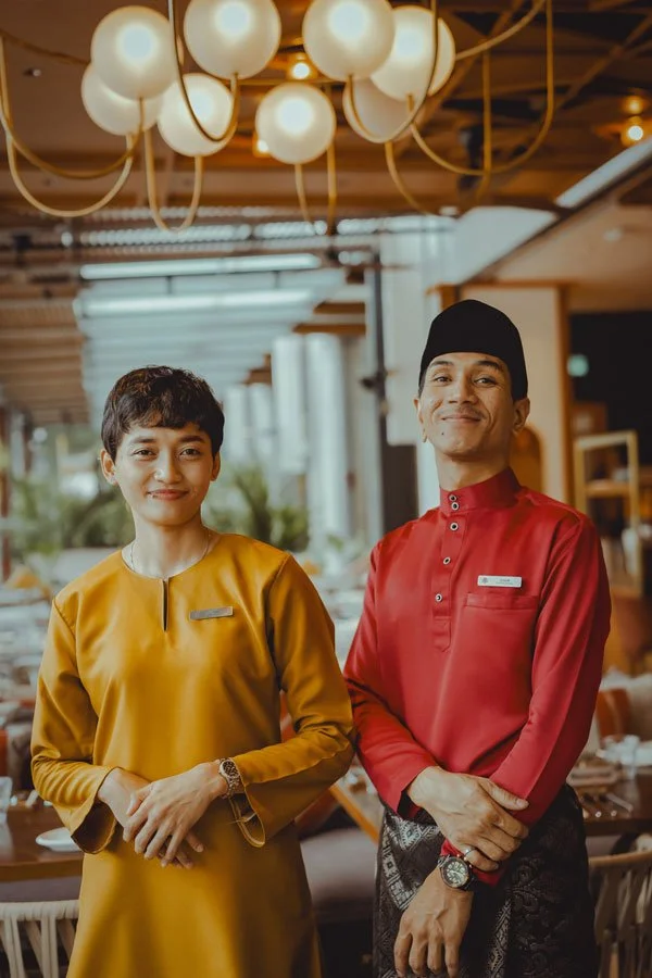 Two restaurant staff members, a woman in a mustard yellow traditional dress and a man in a red traditional outfit with a black hat, standing inside a restaurant with warm lighting and modern decor.