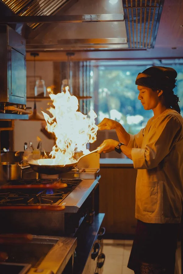 A chef in a black head covering and beige coat cooks on a stove with a large flame in a professional kitchen.