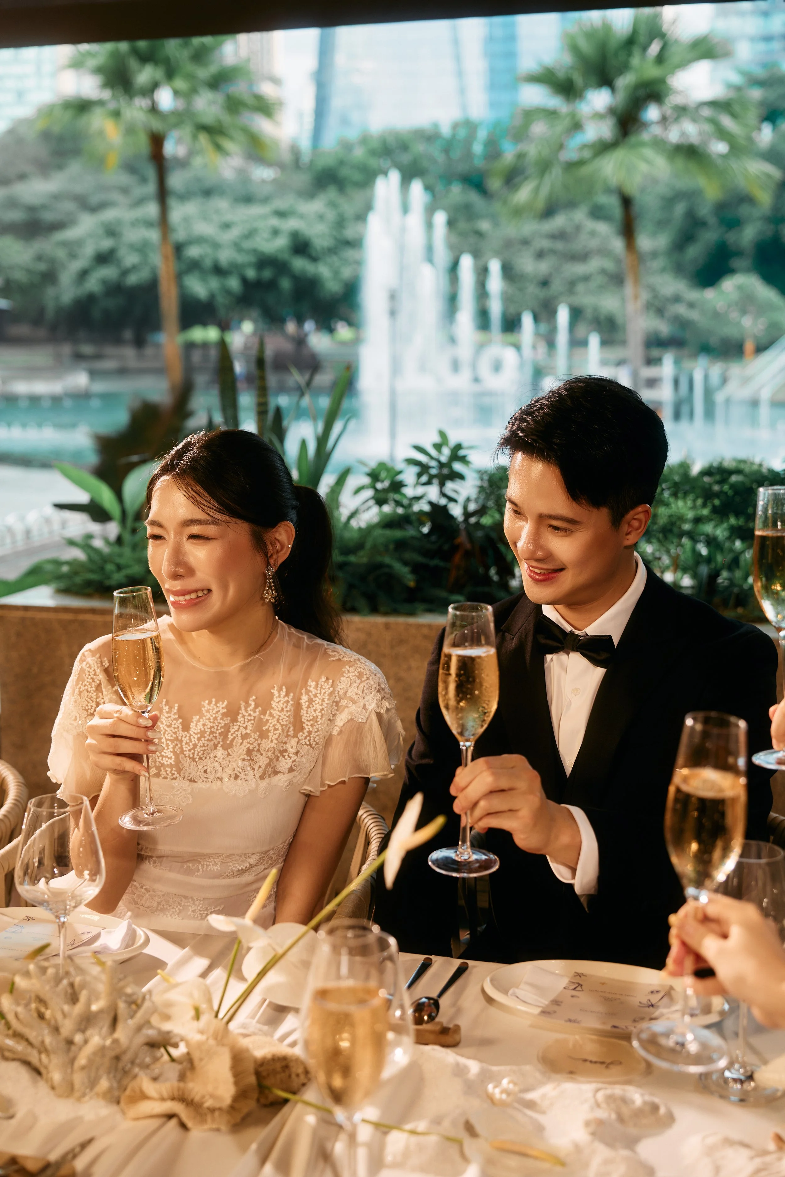 A bride and groom celebrating at a wedding reception, holding champagne glasses, seated at a decorated table with flowers, in an indoor venue with a view of a fountain and greenery outside.