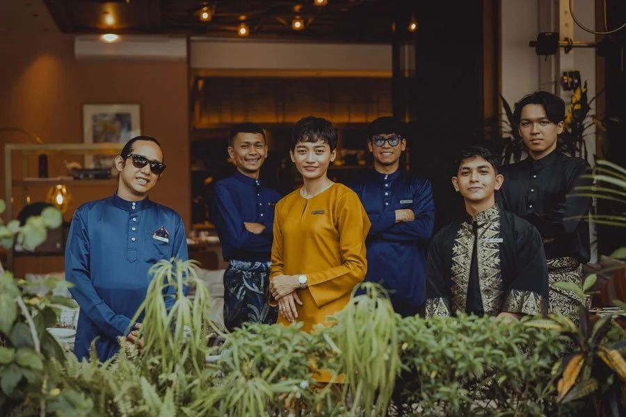 Six people dressed in traditional and modern ethnic attire standing behind a garden of green plants in a restaurant or cafe setting.