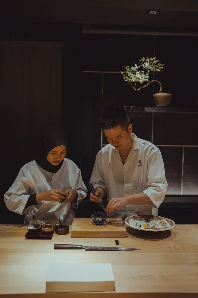 Two chefs in white uniforms preparing food behind a wooden counter in a dimly lit Japanese restaurant.