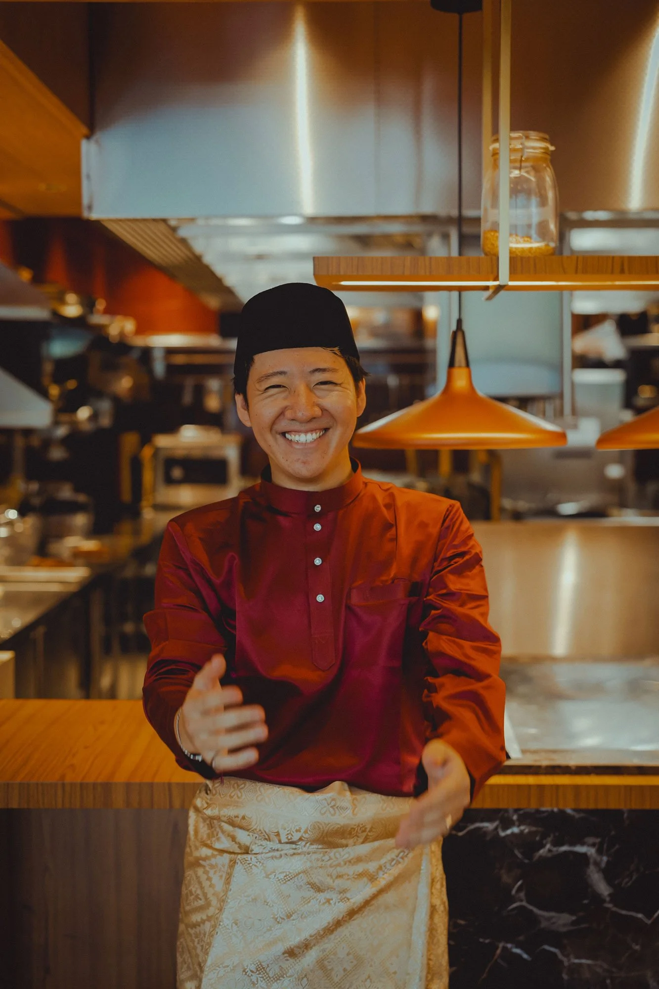 A smiling Thai chef wearing a traditional chef uniform with a red shirt, black hat, and beige apron standing in a restaurant kitchen.