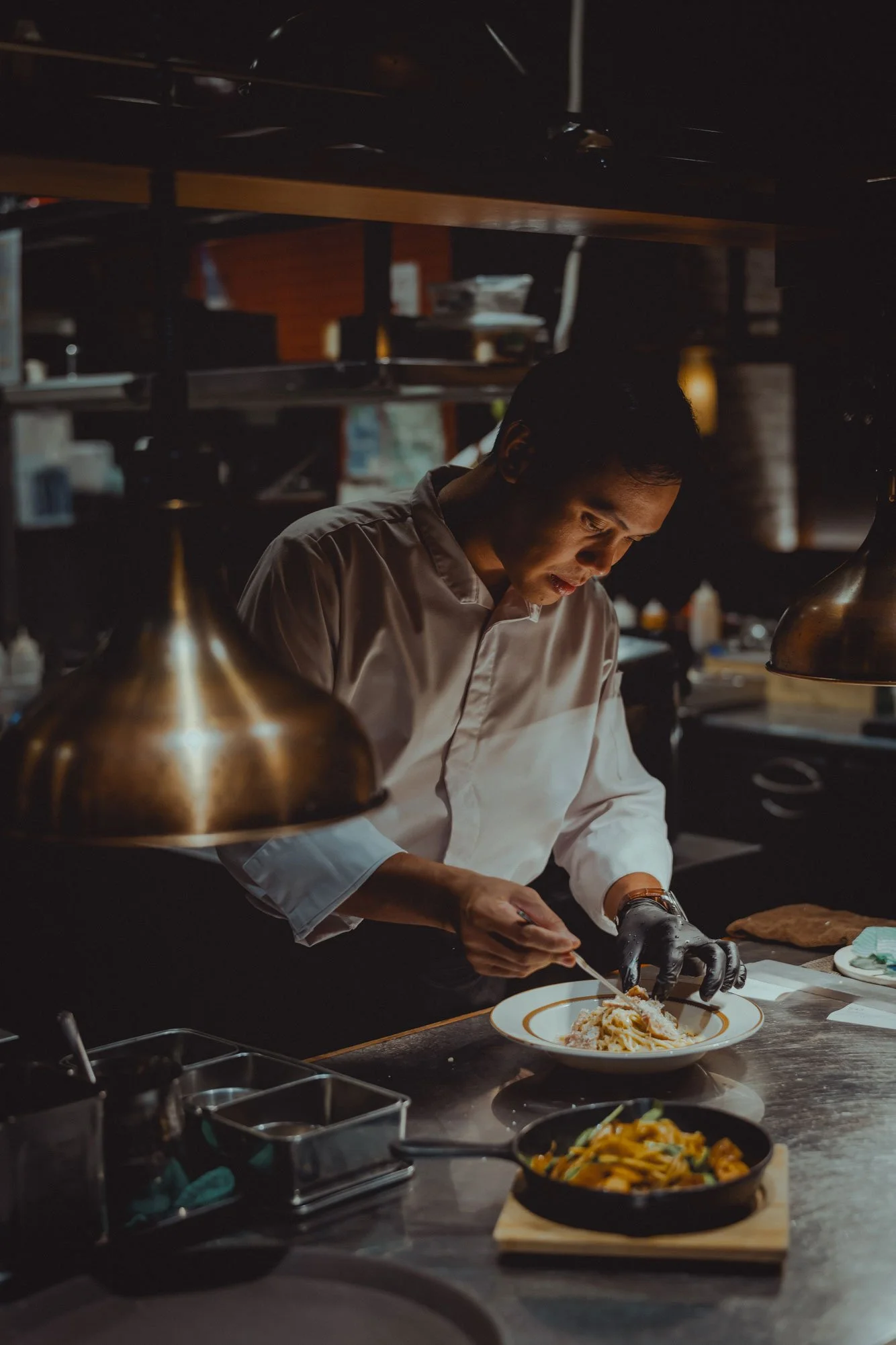 A chef in a professional kitchen plating a dish of pasta, with another pan of pasta on the counter.