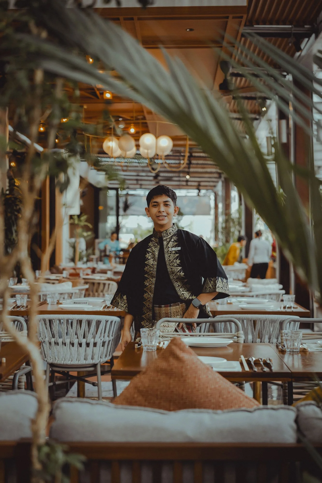 A young man in traditional attire standing inside a restaurant with set tables, glassware, and a cozy, well-lit interior.