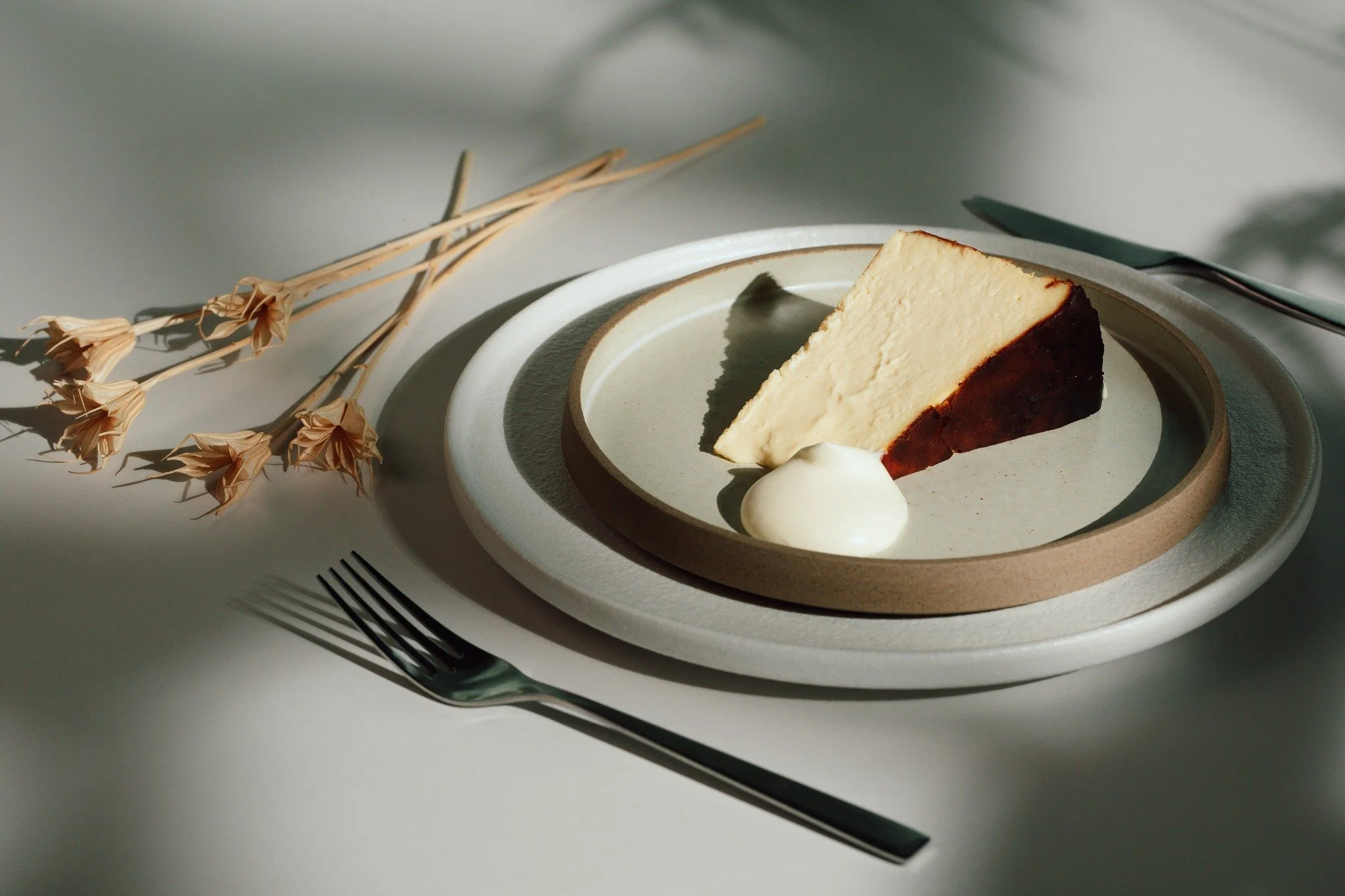 A slice of cheesecake on a plate with a dollop of cream, set on a minimalist table with a fork, knife, and dried flowers nearby.
