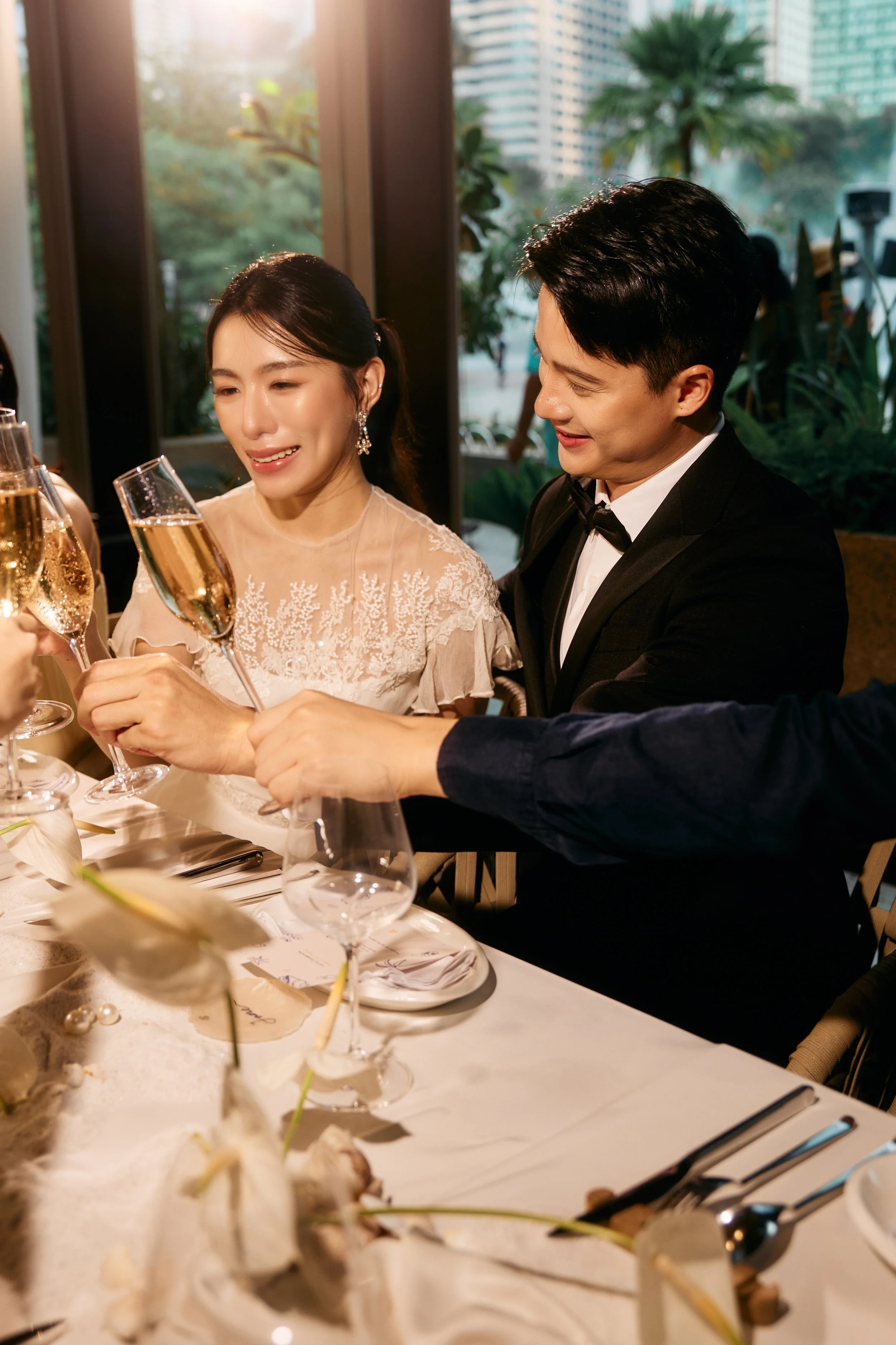 A group of people at a formal event, raising champagne glasses in a toast, with a woman in a white lace dress and a man in a tuxedo.