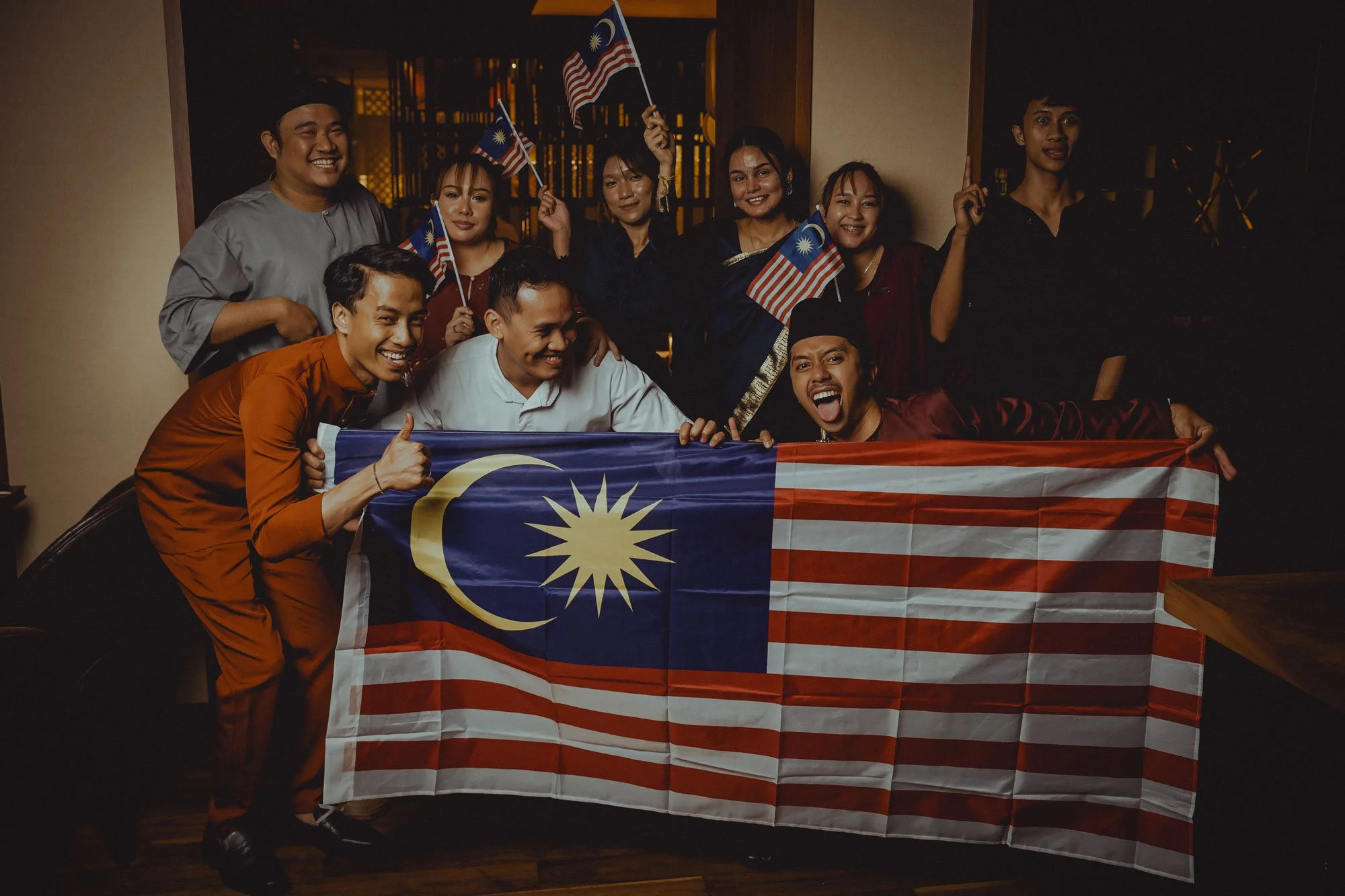 Group of people celebrating Malaysia Independence Day, holding Malaysian flags and draping a Malaysian flag in front.