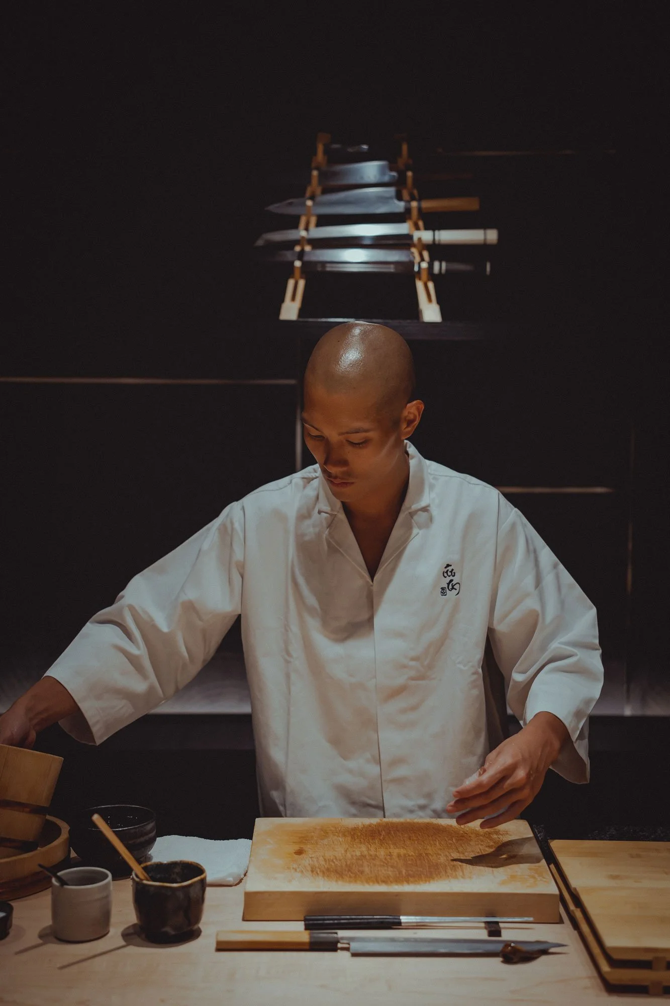 A chef in a white uniform preparing food on a wooden cutting board in a dimly lit kitchen with Japanese knives and utensils.