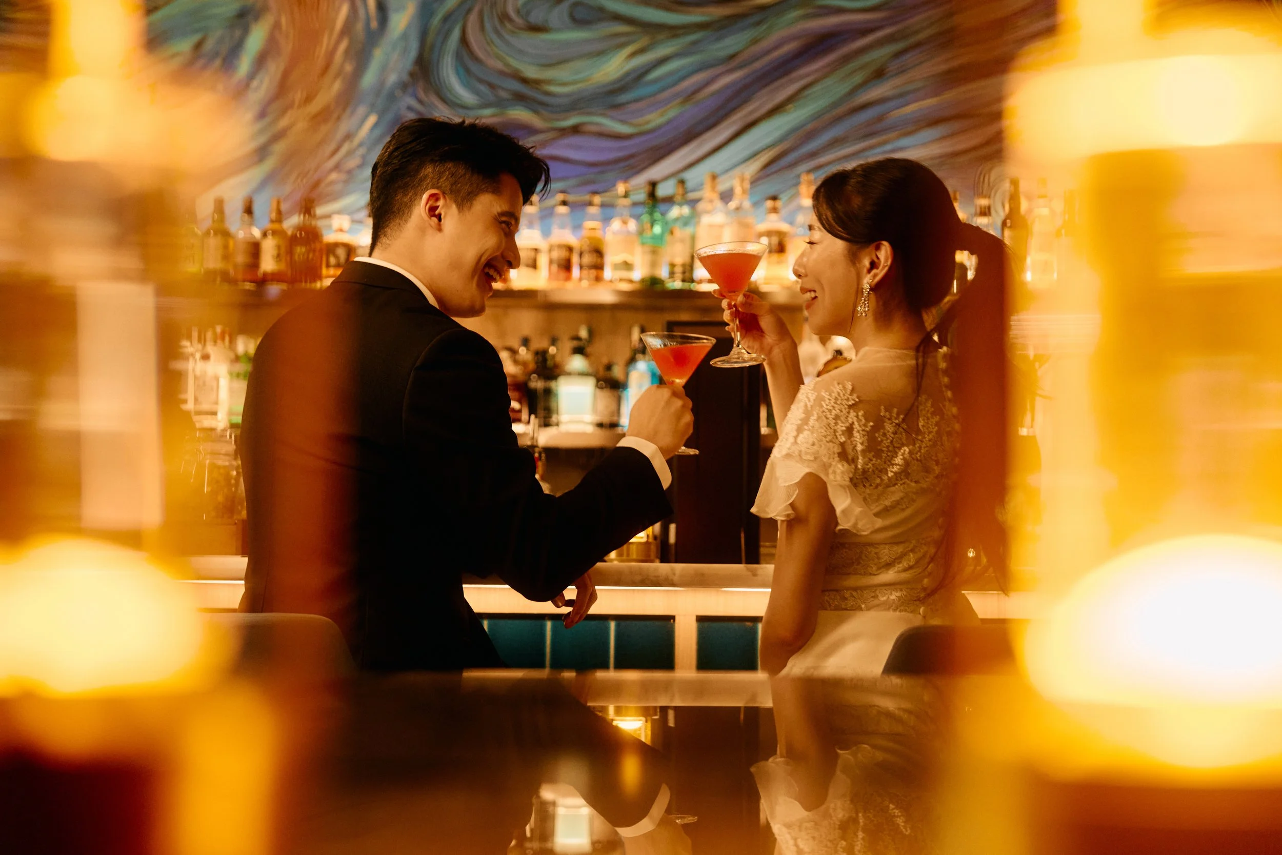 A man and woman in formal attire sharing a toast with cocktails in a dimly lit bar with colorful artwork on the wall.